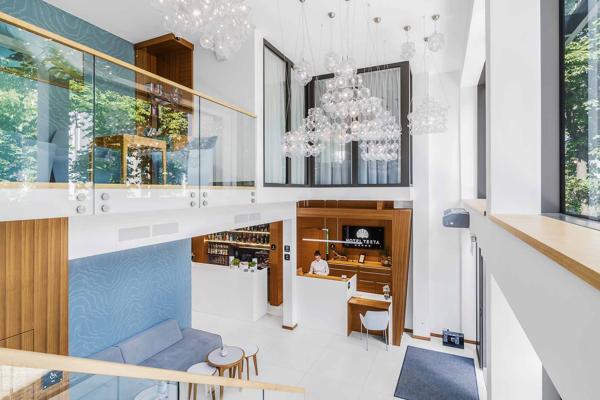 Nowoczesne lobby hotelowe z wysokim sufitem i dużymi oknami zapewnia zachęcającą atmosferę, idealną do sesje fotograficzne wnętrz Gdańsk Gdynia Sopot – usługi fotograficzne. Po prawej stronie widać recepcję z pracownikiem, a po lewej przytulny kącik wypoczynkowy z niebieską sofą. Antresola ze szklanymi balustradami i wiszącymi lampami