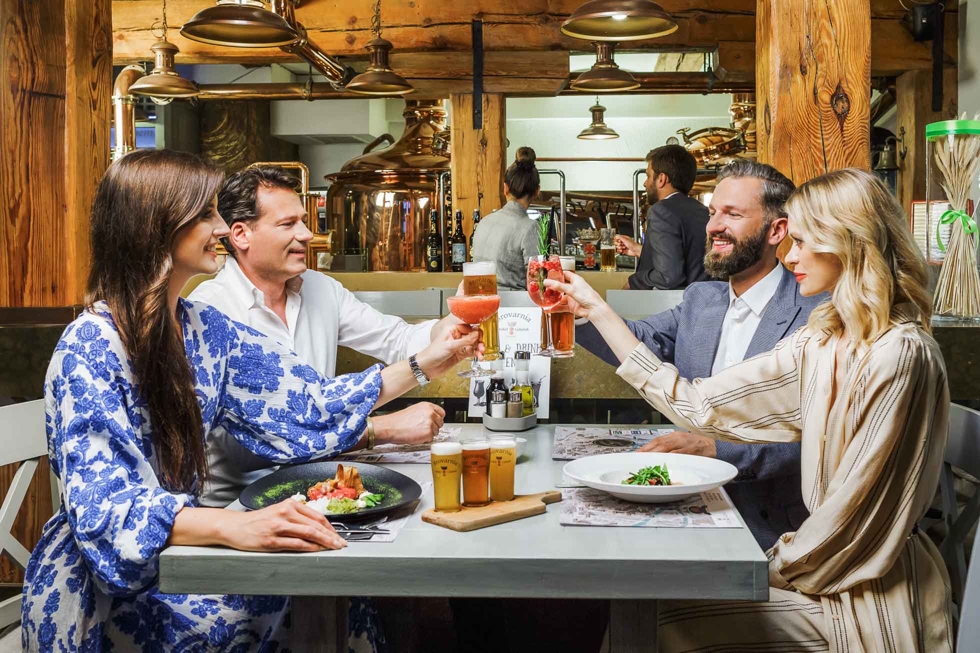 Cztery osoby siedzą przy stoliku w restauracji, wznosząc toast drinkami. Stół jest ozdobiony talerzami z jedzeniem i napojami, pokazującą sesje wizerunkowe dla restauracji i hoteli Gdańsk. Rustykalne wnętrze z drewnianymi belkami i kadziami browaru