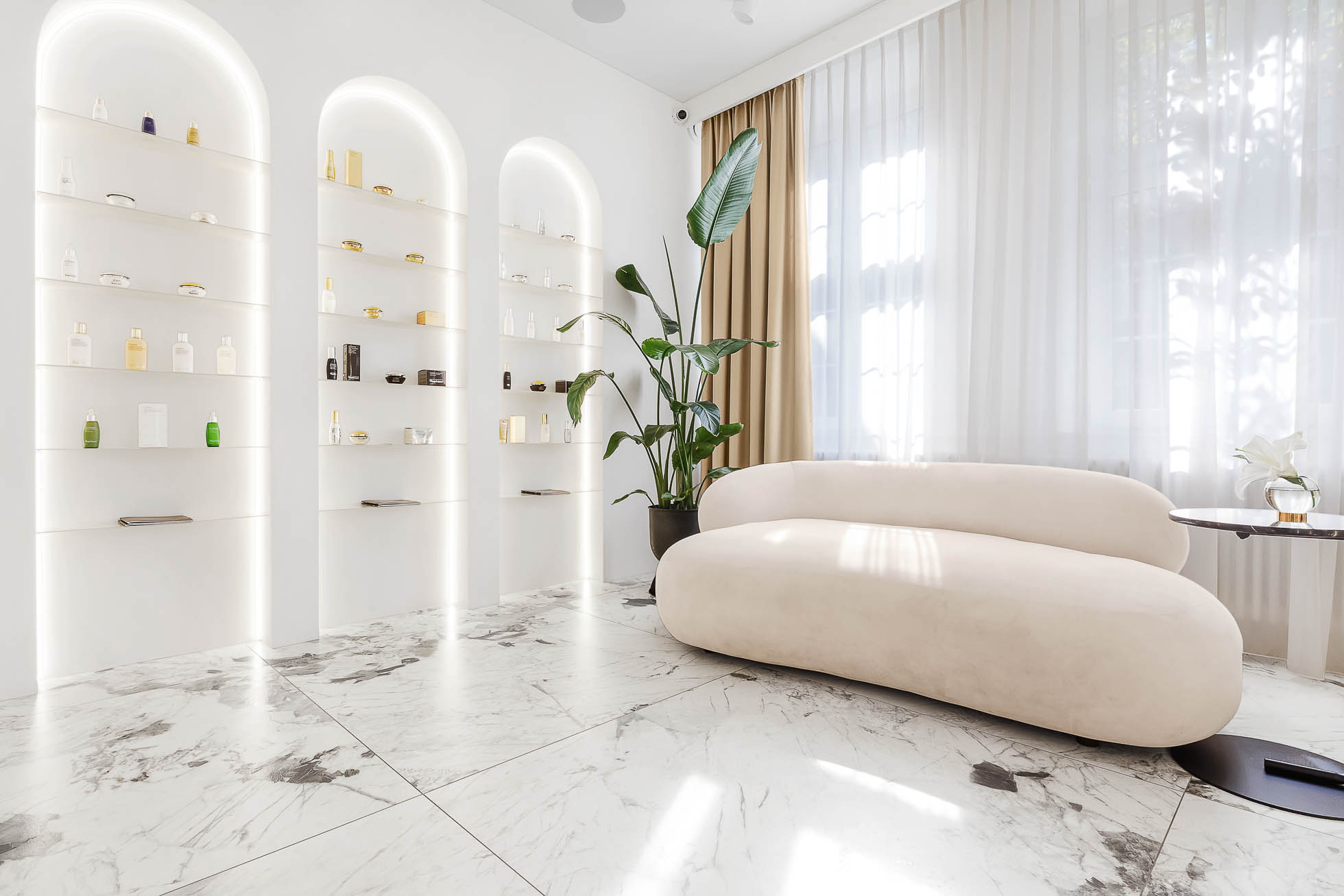 Nowoczesny, minimalistyczny salon z zakrzywioną beżową sofą na marmurowej podłodze. Półki z produktami do pielęgnacji skóry są oświetlone w trzech zagłębionych łukach na lewej ścianie. Wysoka zielona roślina i miękkie naturalne światło z dużych okien z cienkimi firankami dodają spokojnej atmosfery, idealnej do fotografia architektury Trójmiasto – zdjęcia wnętrz i budynków.
