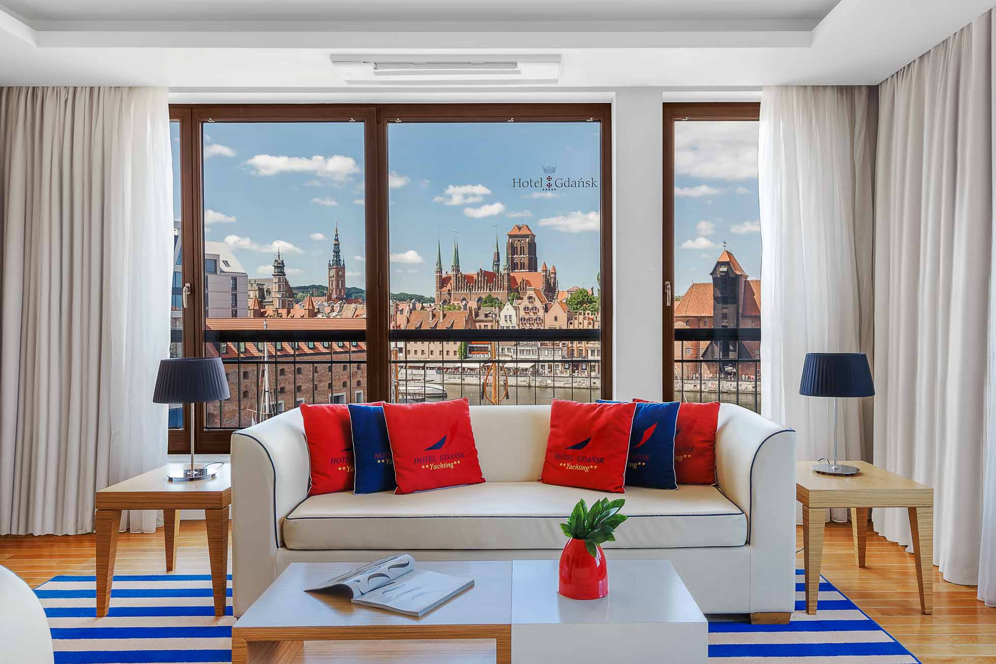 Jasny pokój hotelowy z białą kanapą z czerwonymi i niebieskimi poduszkami z napisem „Hotel Gdańsk”. Stolik kawowy mieści czasopismo i roślinę. Duże okna eksponują zabytkowe budynki Gdańska pod błękitnym niebem, idealne dla Fotografia wnętrz Trójmiasto – zdjęcia apartamentów i hoteli.