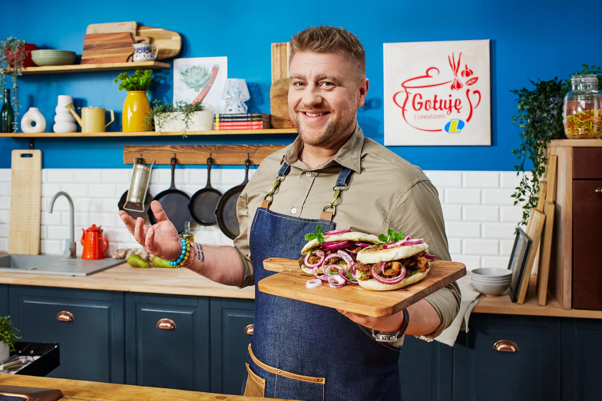 Uśmiechnięty Tomasz Jakubiak w kuchni trzyma dwie drewniane deski, na każdej z nich kanapki. Kuchnia ma niebieskie szafki, wiszące naczynia kuchenne i różne przedmioty. Na ścianie widnieje napis „Gotuje z lewiatanem”. Zdjęcie wykonała Fotografia reklamowa i kulinarna Gdańsk.