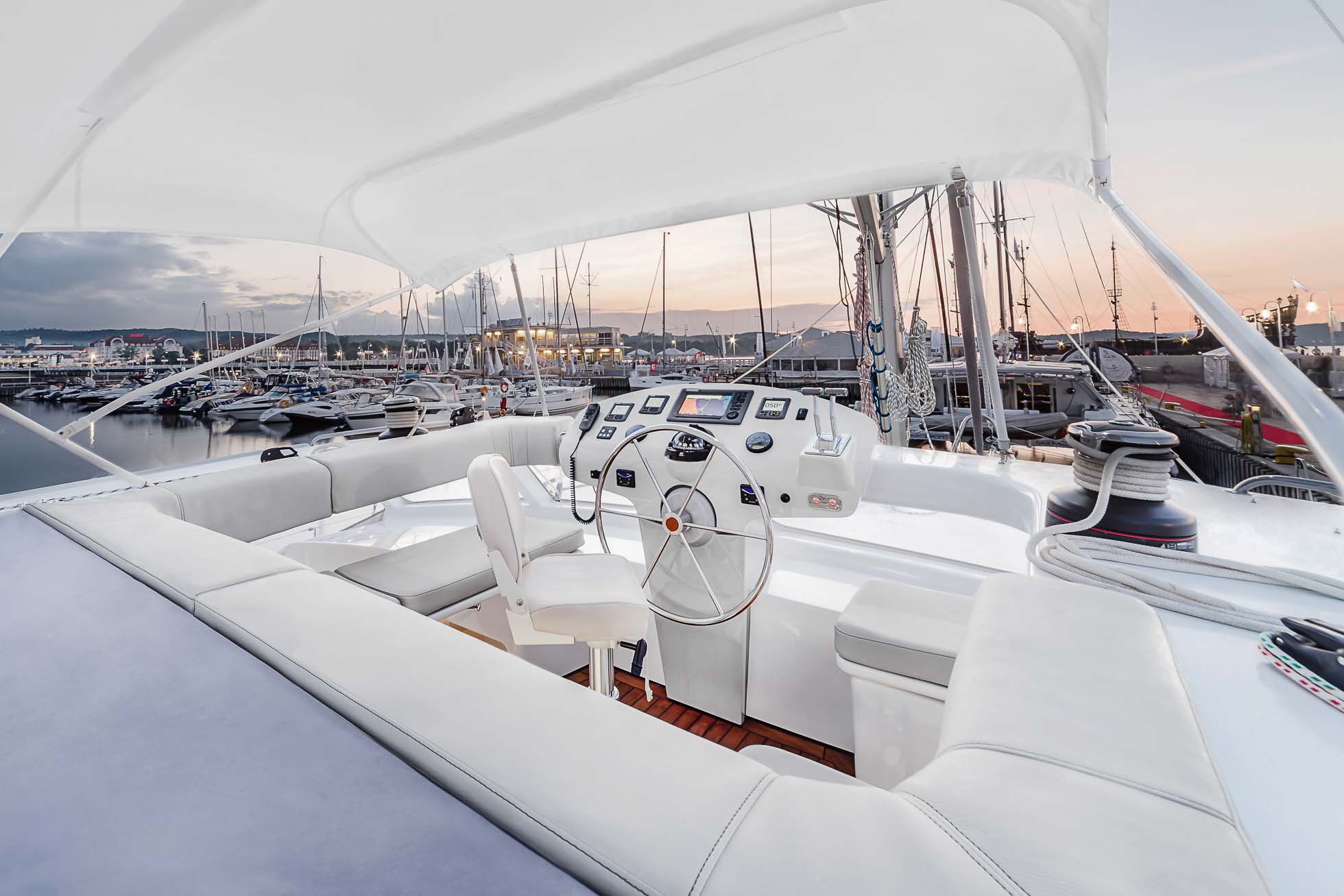 Elegancki, nowoczesny pokład jachtu z białym fotelem i kołem sterowym. Wygodne białe siedzenia otaczają obszar. Jacht jest zacumowany w marinie na końcu molo w Sopocie, z widocznymi innymi łodziami i zachodem słońca w tle — idealne miejsce na profesjonalną fotografię jachtów Trójmiasto.