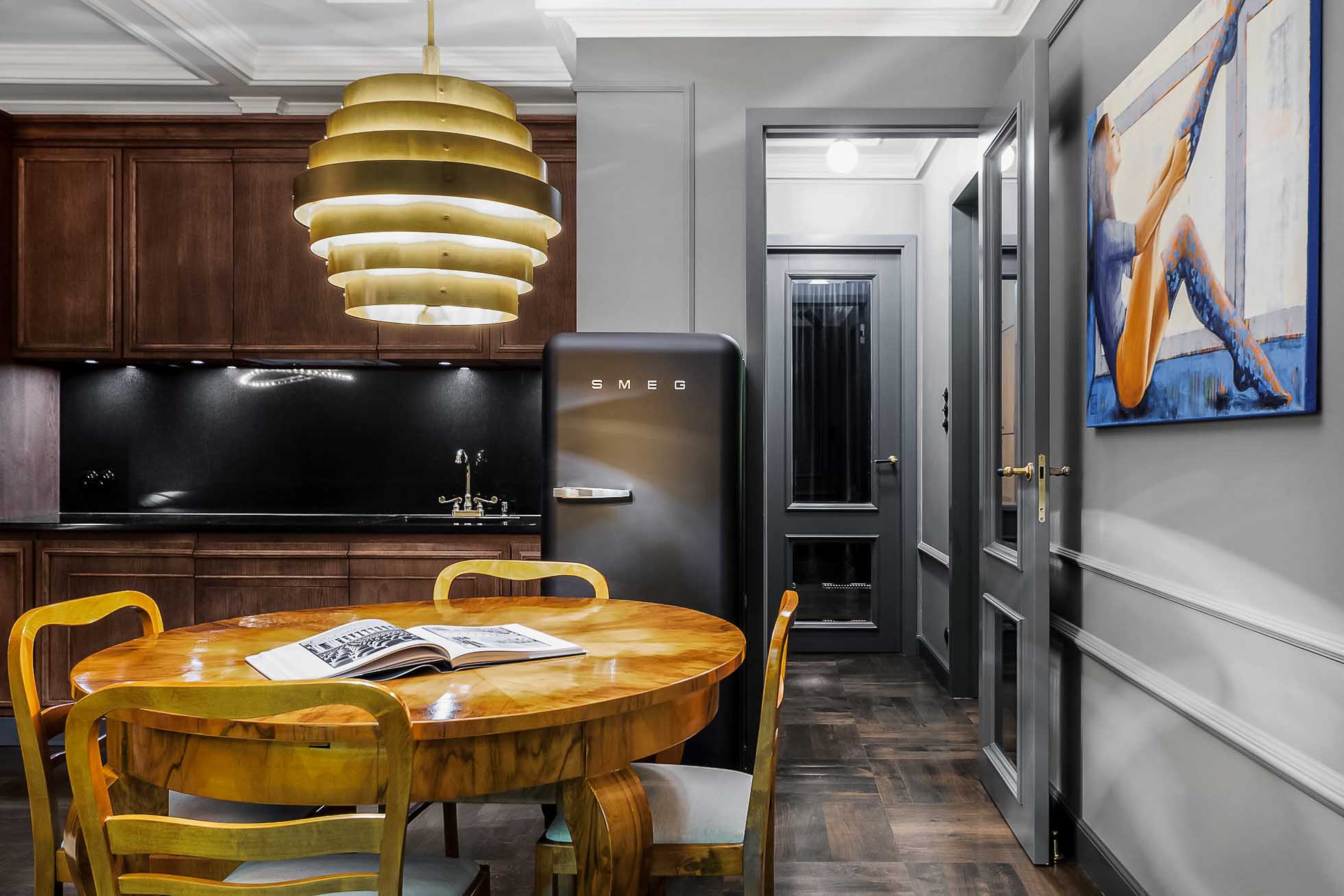 Przytulna kuchnia wyposażona jest w okrągły drewniany stół jadalny z czterema krzesłami. Na stole pod nowoczesną, warstwową lampą wiszącą leży otwarty magazyn. Czarna lodówka Smeg stoi obok drewnianych szafek, a obraz baletnicy zdobi ścianę po prawej stronie. Fotograf wnętrz premium w Gdańsku Gdyni i Sopocie pięknie uchwycił tę przestrzeń.