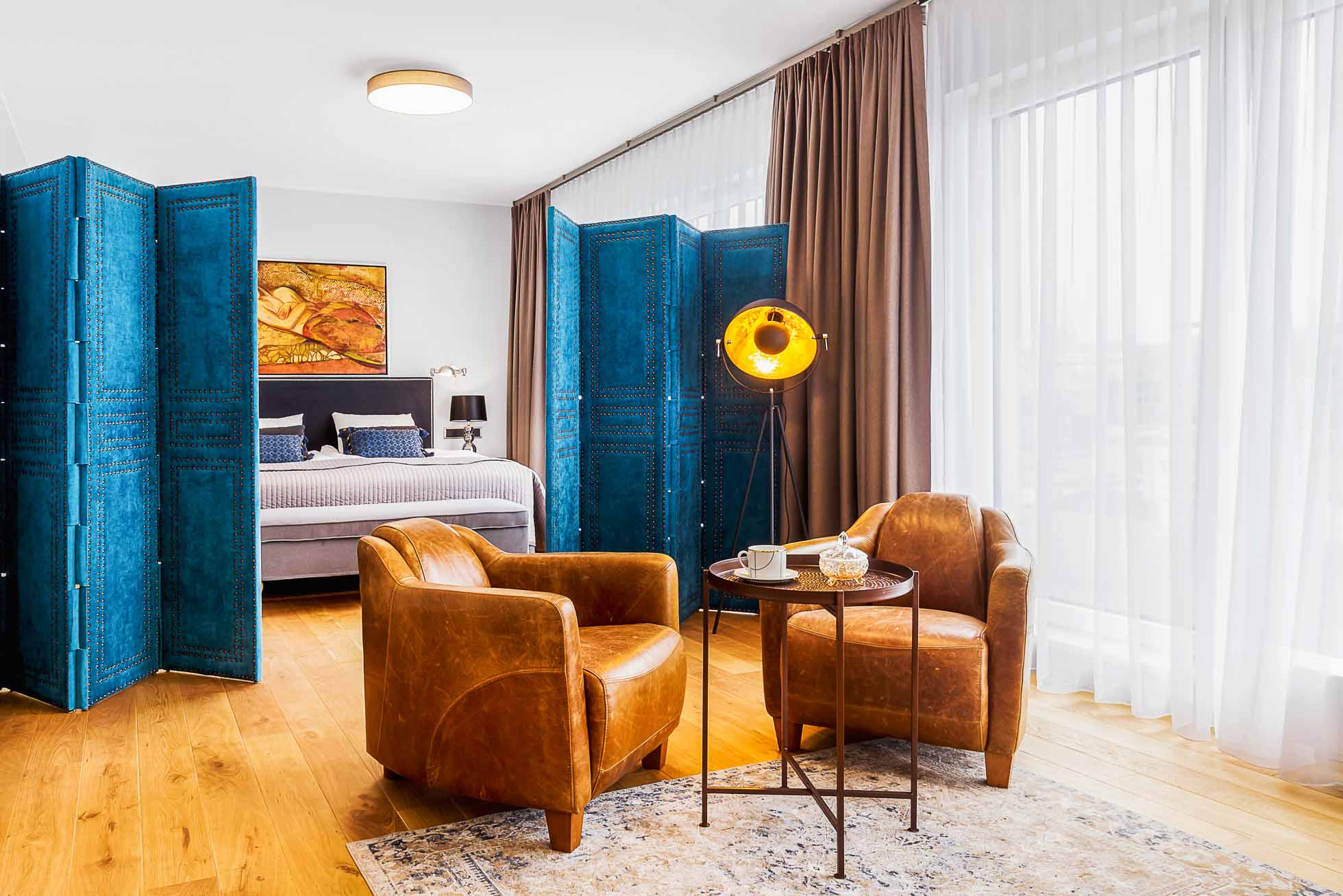 Przytulna przestrzeń życiowa z dwoma brązowymi skórzanymi fotelami wokół małego okrągłego stołu z kubkiem i książkami. Niebieskie ścianki działowe oddzielają sypialnię od łóżka i dzieł sztuki nad nim. Przezroczyste białe zasłony zakrywają duże okna, wpuszczając naturalne światło. Fotograf wnętrz premium w Gdańsku Gdyni i Sopocie pięknie uchwycił to spokojne otoczenie.