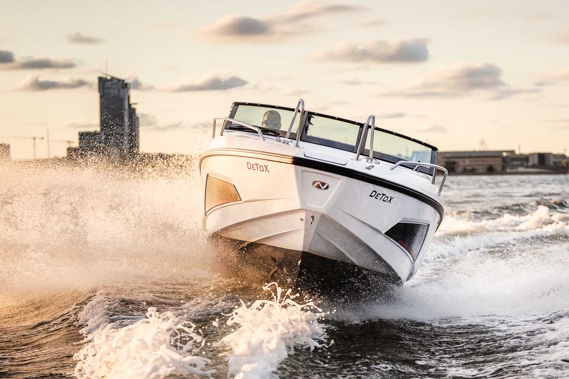 Biała motorówka o nazwie „Detox” muska powierzchnię wody, tworząc ślad podczas szybkiego ruchu do przodu. Słońce jest nisko na niebie, rzucając ciepły blask na scenę. W tle widoczne są budynki miejskie i wysoki wieżowiec, idealne dla Fotografia komercyjna Trójmiasto.