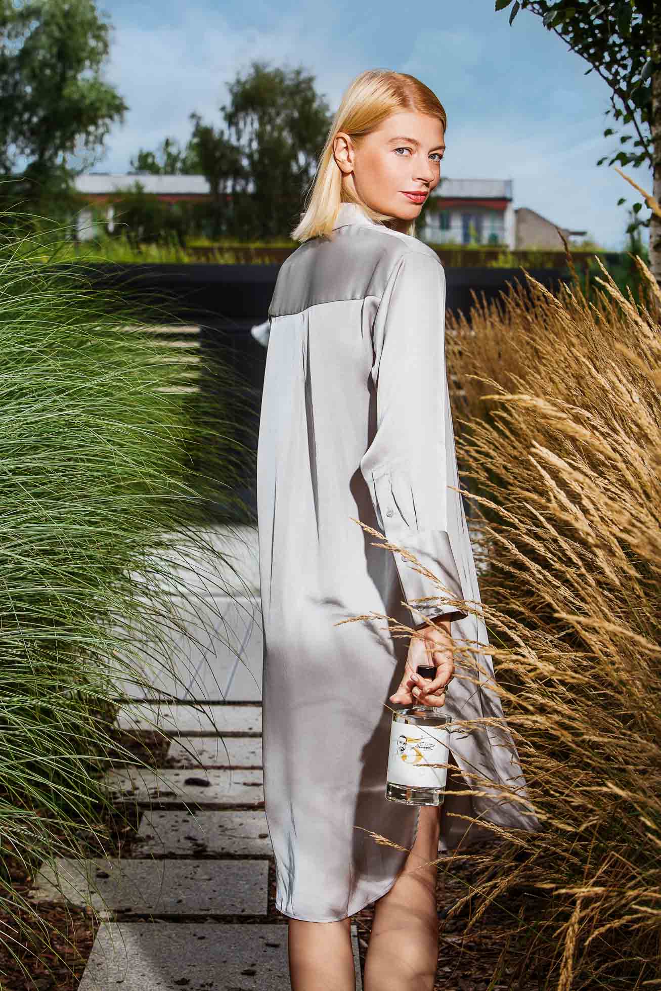 Młoda kobieta o blond włosach stoi na zewnątrz na wybrukowanej ścieżce otoczonej wysoką trawą. Ma na sobie jasnoszarą sukienkę i trzyma w prawej ręce małą, ozdobną butelkę. W tle widać drzewa i nowoczesne budynki pod częściowo zachmurzonym niebem, idealne na profesjonalne kampanie reklamowe Gdańsk.