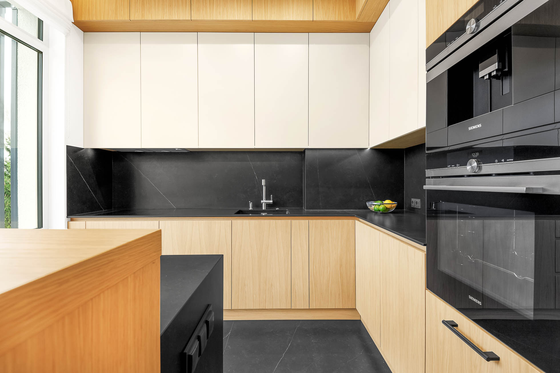 Nowoczesna kuchnia z jasnym drewnem i białymi szafkami, czarnymi blatami i czarnym backsplashem. Wbudowany piekarnik i kuchenka mikrofalowa znajdują się po prawej stronie, a zlew znajduje się pod szafkami na środku. Drewniana wyspa jest częściowo widoczna po lewej stronie. Fotograf wnętrz Trójmiasto – profesjonalna fotografia mieszkań i biur doskonale uchwyciła każdy szczegół.