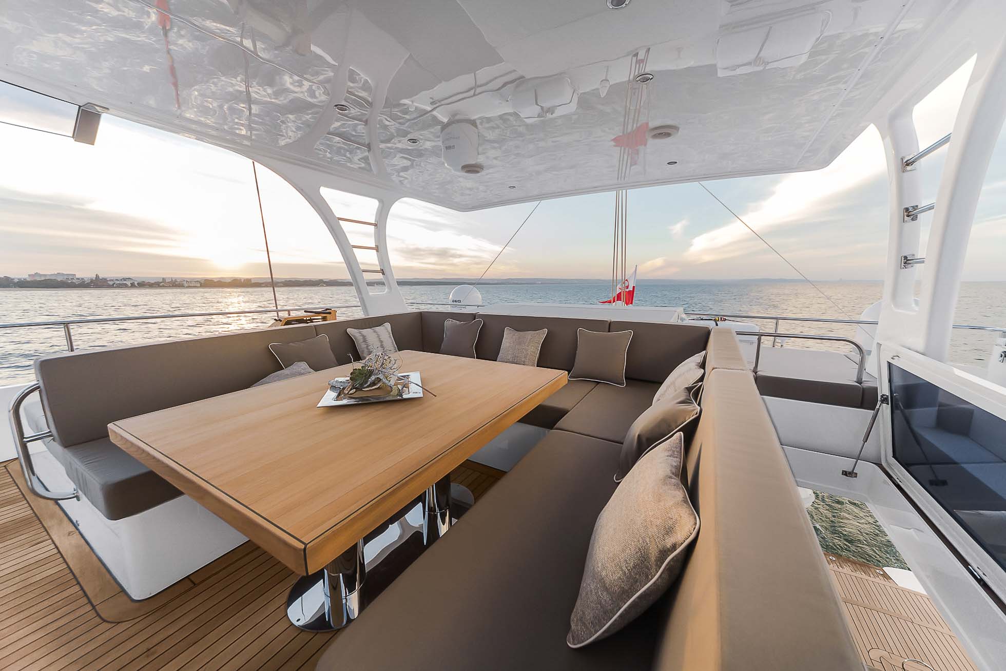 Przestronny pokład jachtu z wyściełanymi siedziskami wokół drewnianego stołu i dekoracyjnej tacy z wazonem i napojami. Część wypoczynkowa oferuje panoramiczny widok na ocean z pięknym zachodem słońca w tle. Elegancki i nowoczesny design, idealny do relaksu. Fotograf wnętrz Trójmiasto – profesjonalna fotografia mieszkań i biur uchwyca te chwile bezbłędnie.