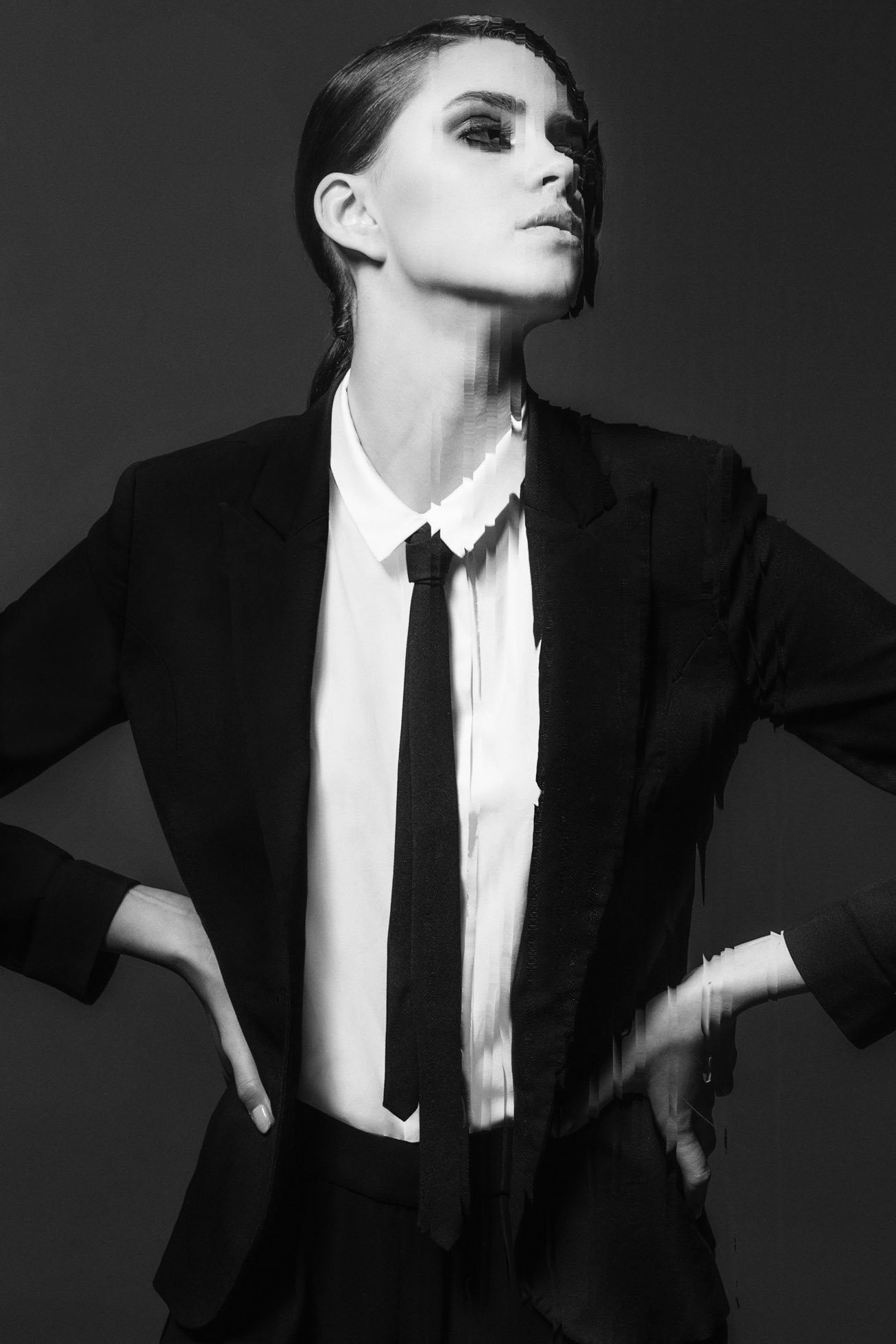 Czarno-biała fotografia wizerunkowa przedstawia osobę w garniturze i krawacie, przyjmującą pewną pozę z rękami na biodrach. Obraz charakteryzuje się intrygującym efektem glitch, dodającym ostry akcent przypominający modowe style edytorial.