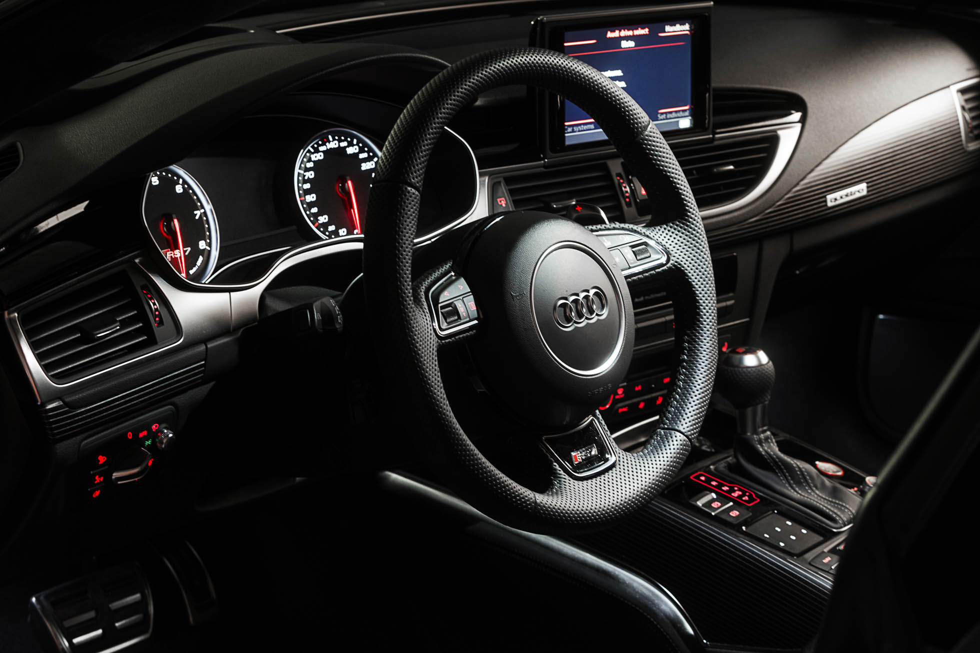 Zbliżenie wnętrza samochodu Audi prezentujące elegancki, czarny design. Widoczna jest kierownica z logo Audi, deska rozdzielcza z prędkościomierzem i obrotomierzem, centralny system sterowania z ekranem, dźwignia zmiany biegów oraz różne przyciski i elementy sterujące. Idealne dla fotografia reklamowa Trójmiasto.