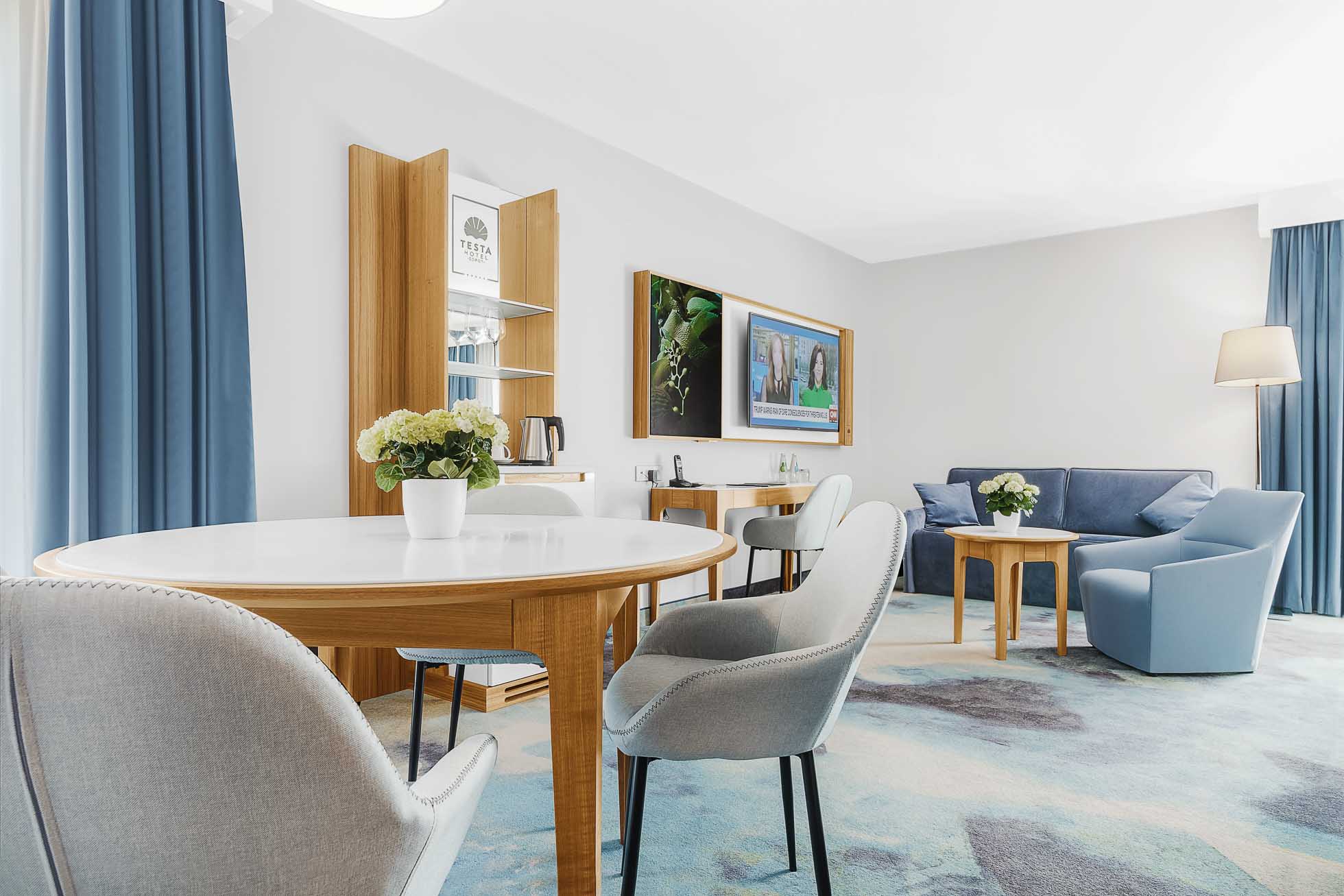 Nowoczesny pokój hotelowy ma okrągły biały stół z drewnianymi nogami, otoczony czterema jasnoszarymi krzesłami. Szafka, telewizor na ścianie, niebieska sofa i mały drewniany stolik z lampą tworzą wygodną strefę wypoczynkową. Niebieskie zasłony i neutralna kolorystyka dodają wystroju. Aby uzyskać nieskazitelne ujęcia takich przestrzeni, rozważ profesjonalną fotografię wnętrz Trójmiasto – najwyższą jakość