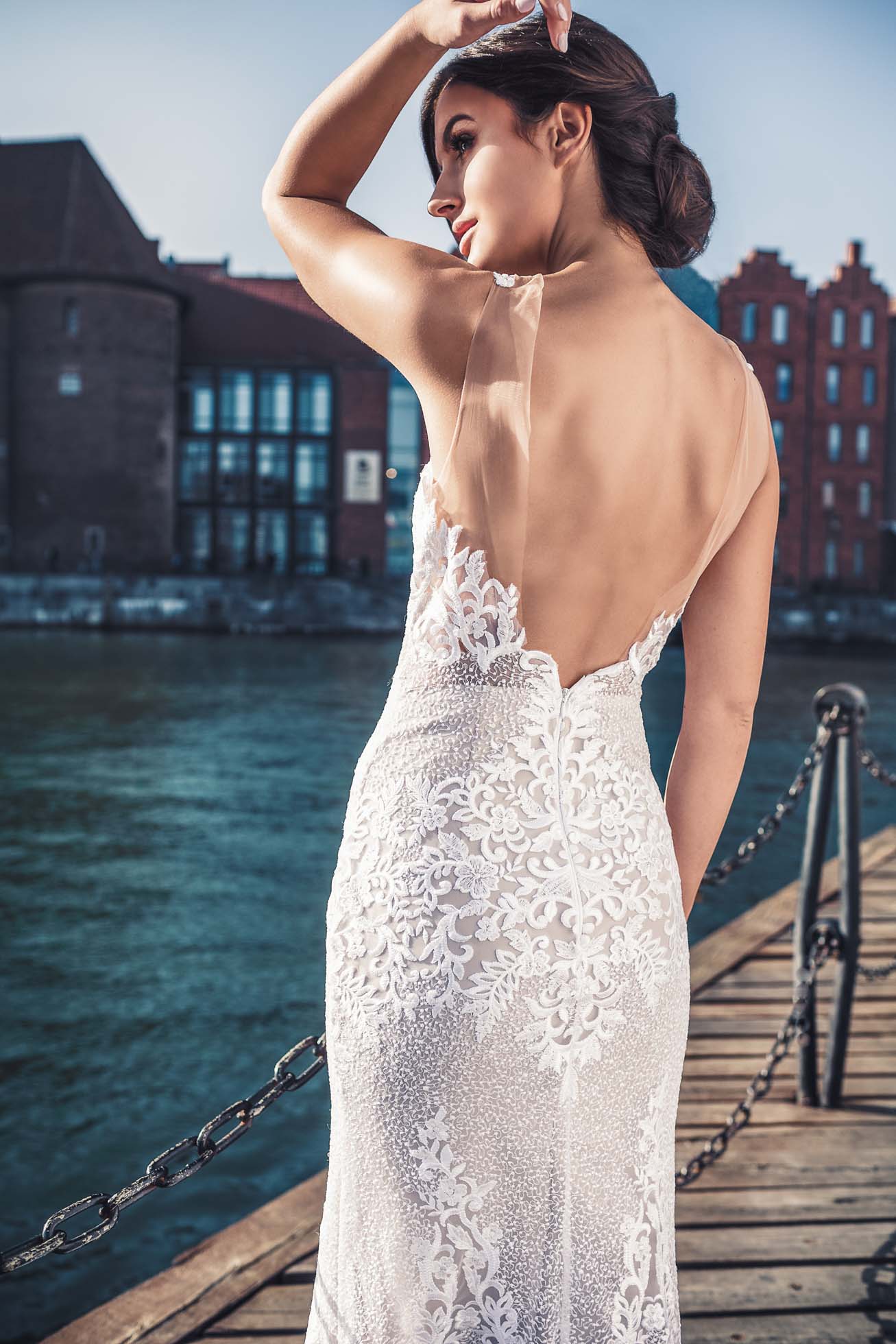 Kobieta w koronkowej sukni ślubnej z głębokim dekoltem stoi na drewnianym molo nad wodą, uchwytując istotę spokojnej elegancji Sopotu. Odwraca wzrok, trzymając się za ręce, podczas gdy ceglane budynki i czyste niebo stanowią malownicze tło godne edytorialu modowego.