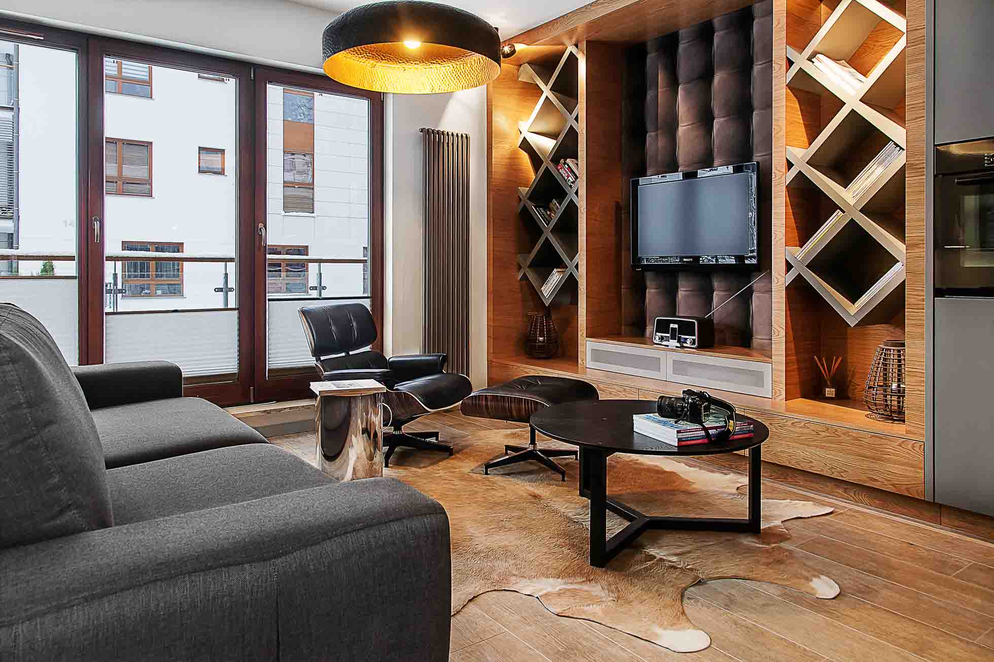 Nowoczesny salon z dużymi oknami, szarą sofą, czarnym fotelem, okrągłym stolikiem kawowym na dywanie z krowiej skóry i telewizorem na ścianie w drewnianym regale z dekoracyjnymi przedmiotami. W pokoju znajduje się ciepłe oświetlenie i nowoczesne wyposażenie. Sesje fotograficzne wnętrz Gdańsk Gdynia Sopot – usługi fotograficzne doskonale oddają jego elegancję.