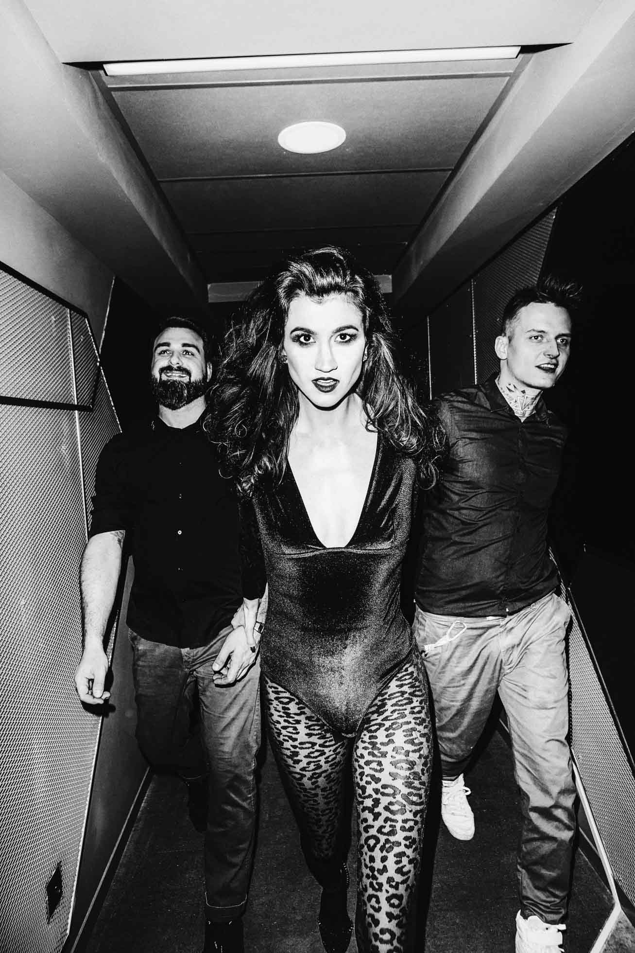 Czarno-biała fotografia wizerunkowa przedstawiająca trzy osoby pewnie idące korytarzem. Centralna postać, ubrana w błyszczący strój ze spodniami w panterkę, emanuje modą edytorialną, podczas gdy osoby po obu stronach noszą swobodne czarne stroje, wszystkie wydają się zaangażowane i dynamiczne.