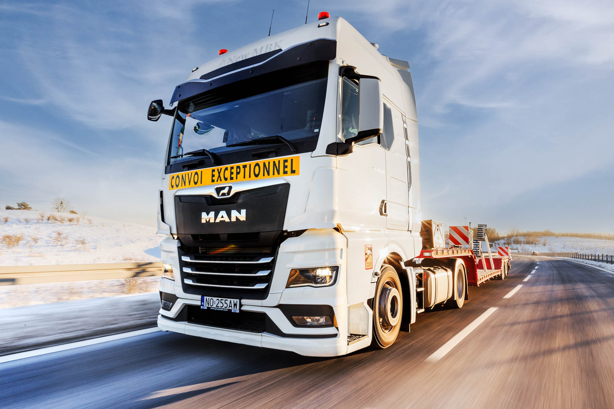 Duża biała ciężarówka MAN z napisem „Convoi Exceptionnel” z przodu jedzie zaśnieżoną autostradą, idealną dla Fotografia biznesowa i korporacyjna Trójmiasto. Ciężarówka ma pustą naczepę z płaską platformą i jest otoczona czystym, błękitnym niebem i śnieżnym krajobrazem.