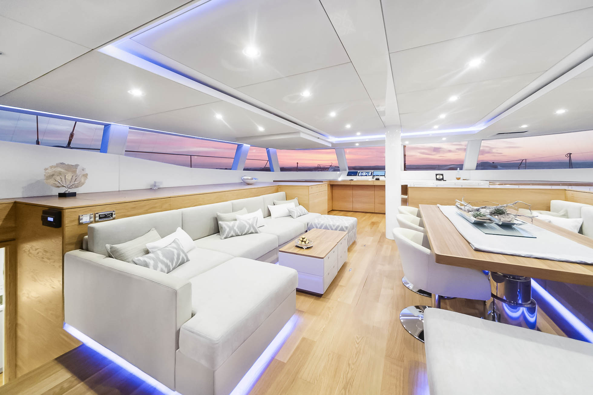Nowoczesne, luksusowe wnętrze jachtu z dużymi oknami, z których widać zachód słońca nad oceanem. Przestrzeń wyposażona jest w białą kanapę modułową, drewniany stolik kawowy, jadalnię z białymi krzesłami i akcenty świetlne w kolorze niebieskim. Uchwycone przez Fotograf wnętrz premium w Gdańsku Gdyni i Sopocie, ogólne wrażenie jest eleganckie i smukłe.