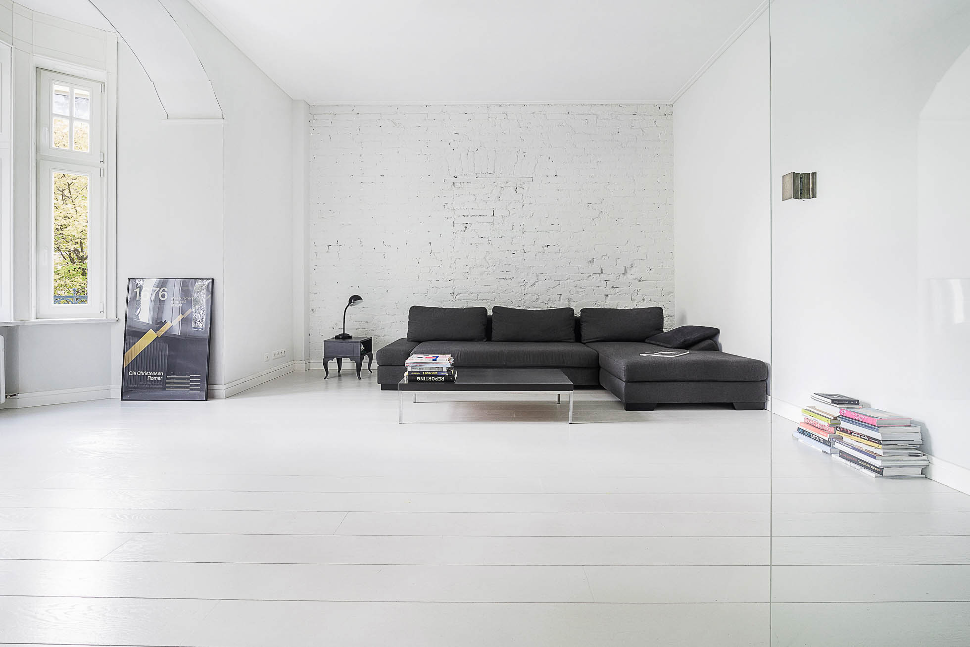 Nowoczesny, minimalistyczny salon z elegancką czarną sofą, stosem książek na przezroczystym stoliku kawowym i lampą na małym stoliku bocznym. Podłoga jest z białych drewnianych desek, a ściany są pomalowane na biało, z czego jedna ma odsłoniętą cegłę. Naturalne światło sączy się przez okno. Ta scena jest idealna dla Profesjonalna fotografia wnętrz Trójmiasto