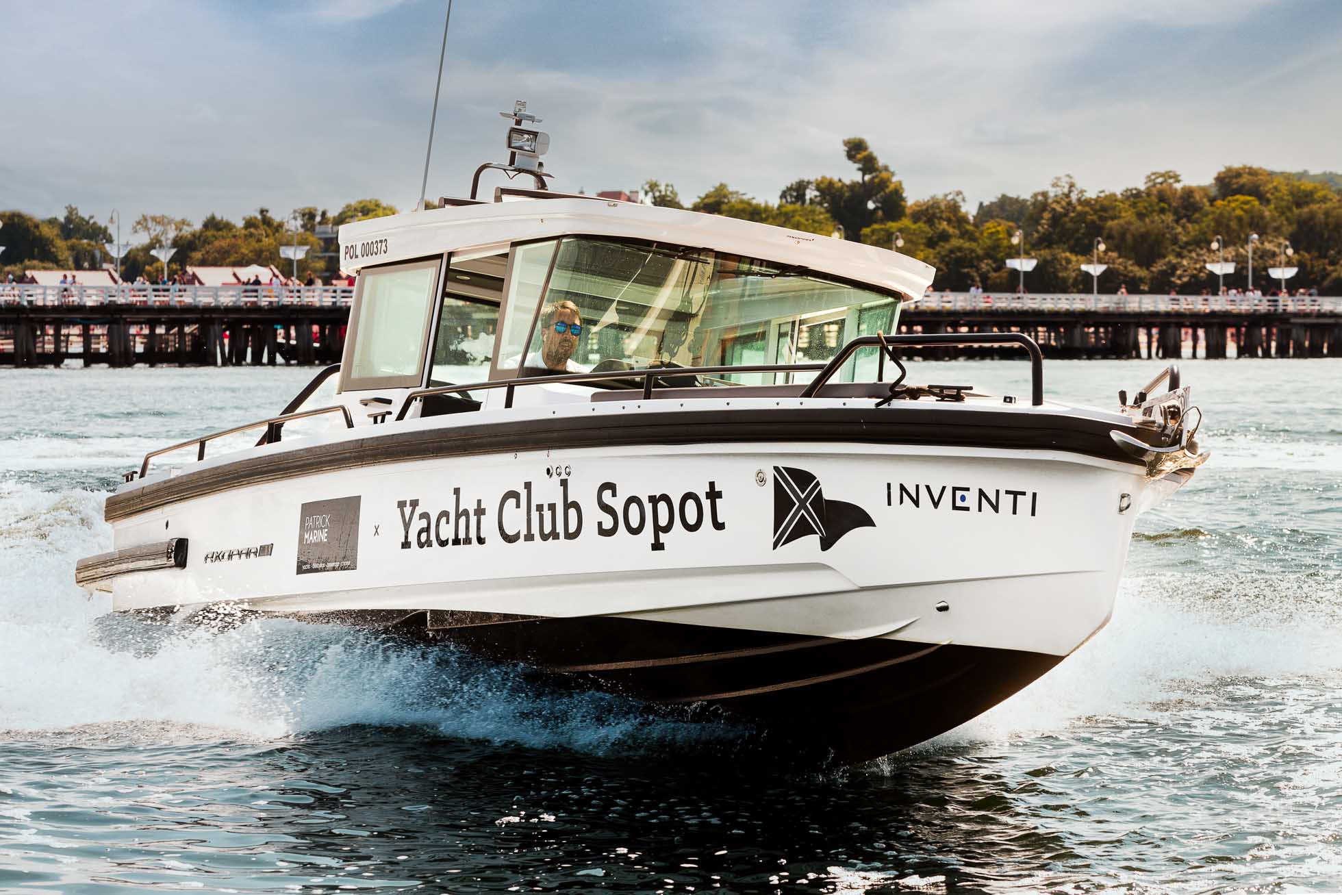 Biała motorówka o nazwie „Yacht Club Sopot” z dodatkowym brandingiem, w tym „Inventi”, żegluje po wodzie. W tle widać pomost i zieleń, gdy łódź tworzy fale, poruszając się do przodu. Wewnątrz kabiny widać osobę sterującą statkiem, idealne do fotografia biznesowa i korporacyjna Trójmiasto.