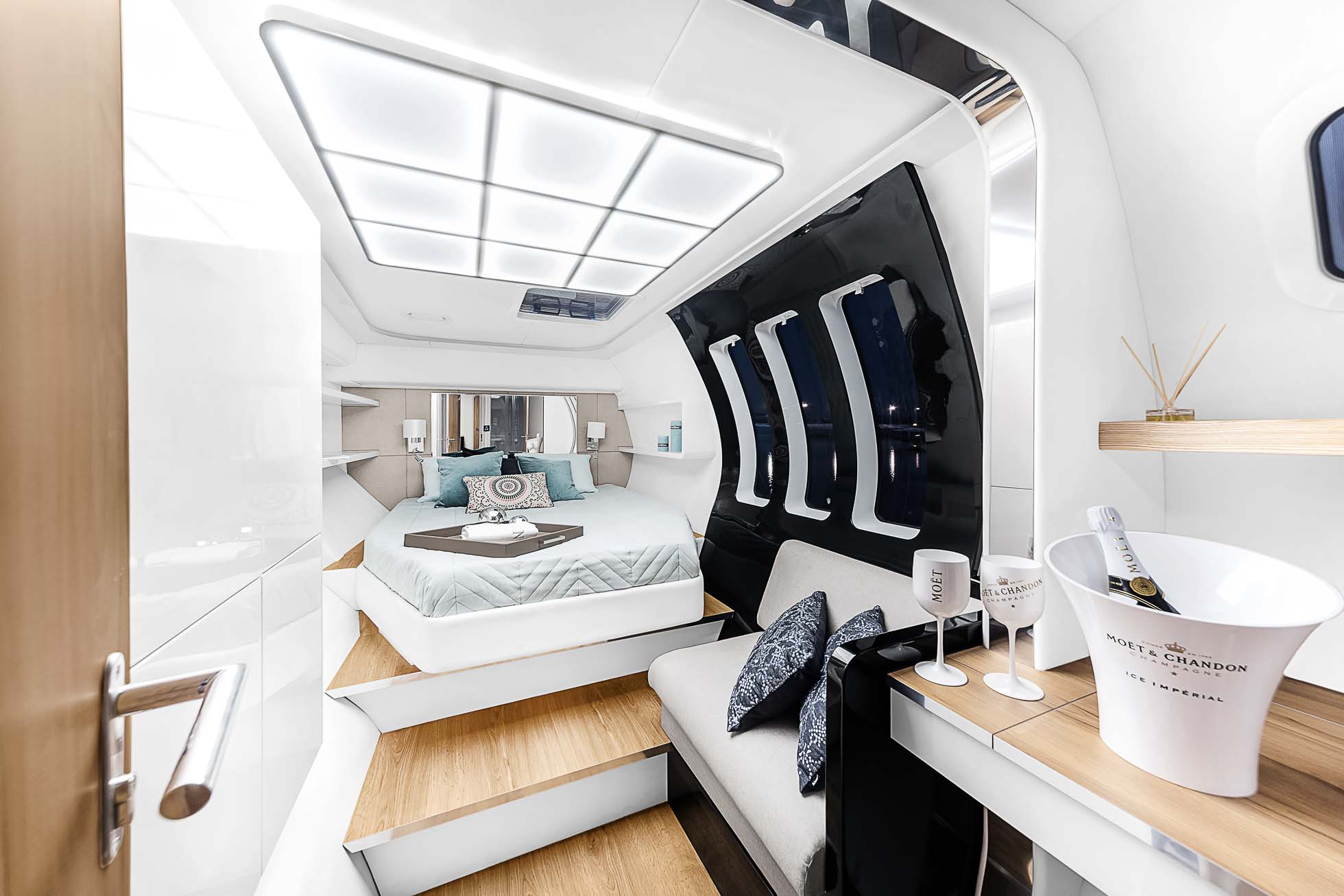 Luksusowa, nowoczesna sypialnia katamaranu z dużym łóżkiem z pastelową pościelą i licznymi poduszkami. Pokój ma gładkie białe ściany, drewniane akcenty i duże czarne lustro. Jest wygodna kanapa, mały stolik z kieliszkami do szampana i butelką, artystyczne oświetlenie na suficie — idealne do sesji Fotografia wnętrz Trójmiasto.