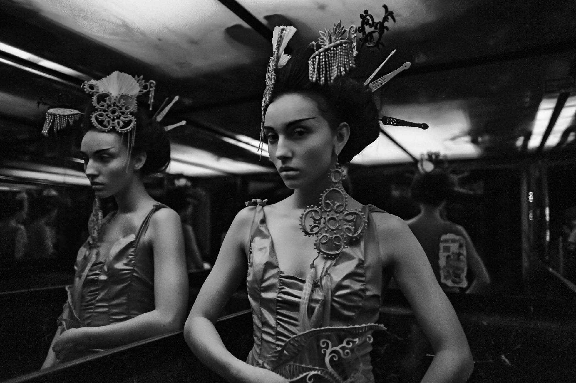 Kobieta w misternej sukni i ozdobnym nakryciu głowy stoi przed lustrem w słabo oświetlonym pokoju, tworząc efekt odbicia. Ta czarno-biała fotografia wizerunkowa podkreśla szczegółowe wzory, uchwytując modową esencję edytorialną z ponadczasową elegancją.