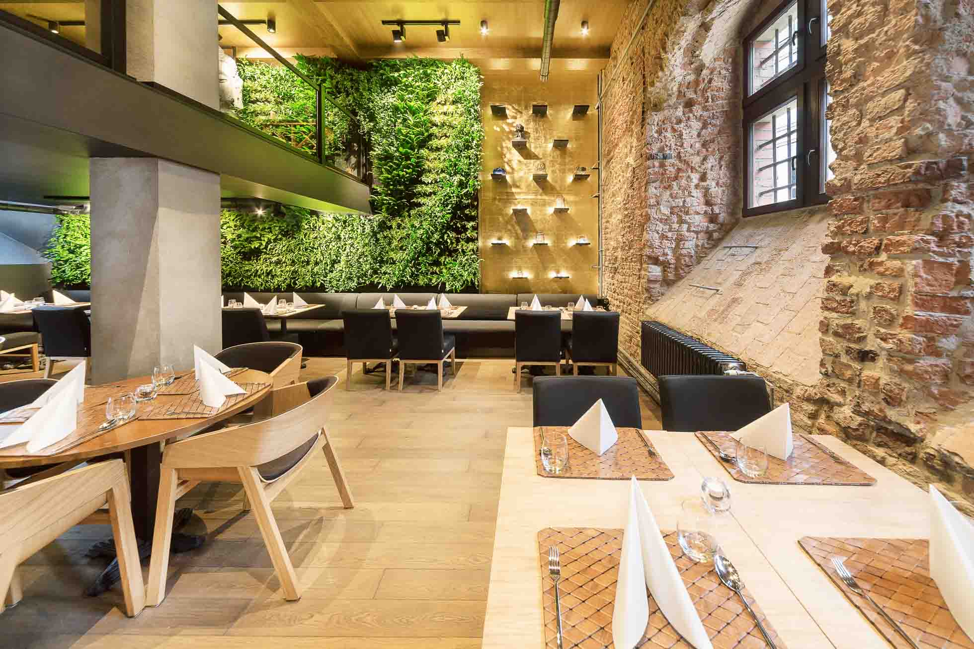 Nowoczesne wnętrze restauracji z pionową zieloną ścianą, odsłoniętymi ścianami z cegły i drewnianymi akcentami. Kilka stolików z tkanymi podkładkami, serwetkami i szklankami jest starannie ułożonych z czarnymi i drewnianymi krzesłami. Duże okna zapewniają naturalne światło — idealne dla Fotograf wnętrz premium w Gdańsku Gdyni i Sopocie.
