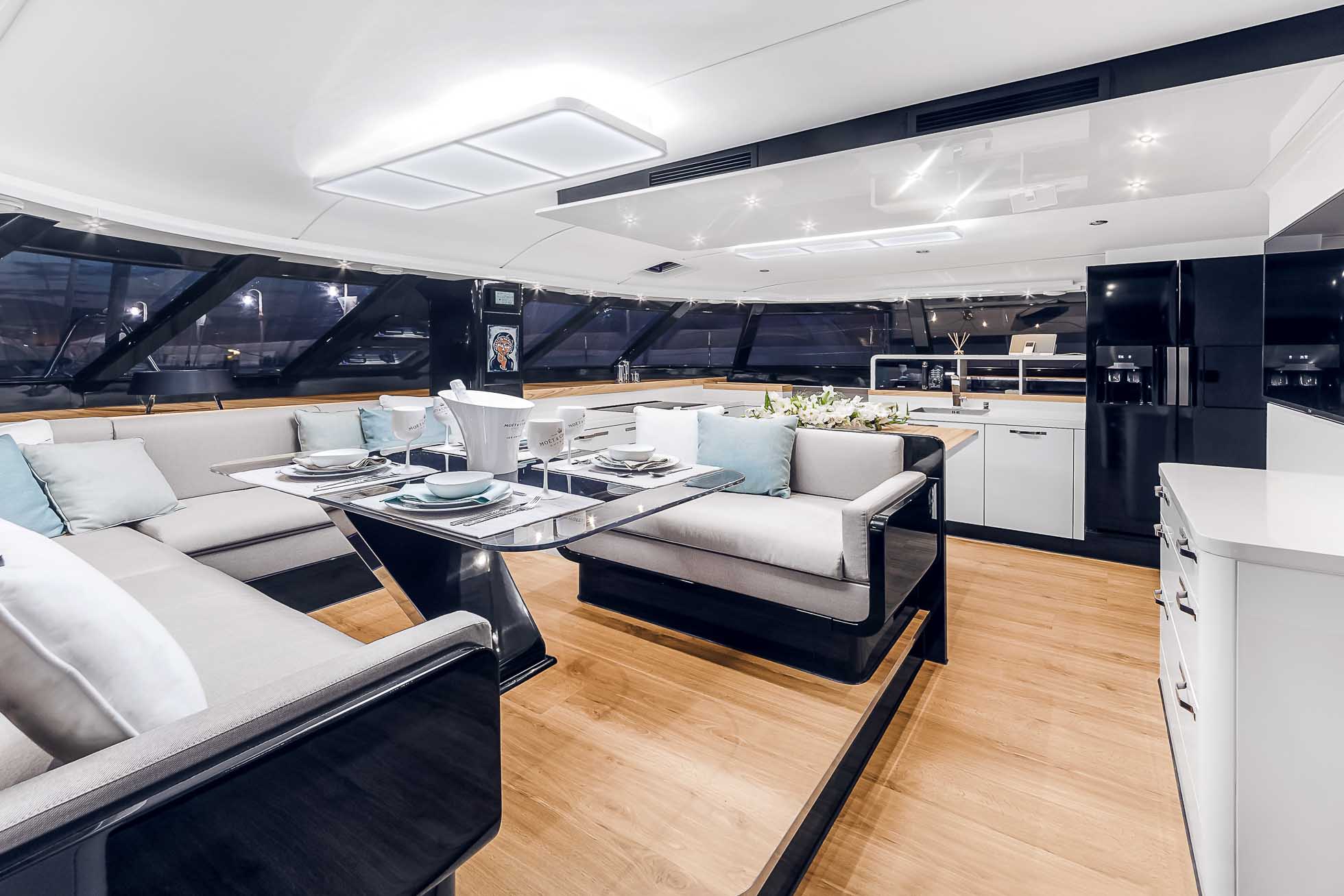 Przestronne, nowoczesne wnętrze jachtu z dobrze oświetlonym miejscem do siedzenia. W pomieszczeniu znajdują się wygodne szare sofy z jasnoniebieskimi poduszkami, elegancki stół jadalny z talerzami i szklankami, drewniana podłoga i duże okna z widokiem na zewnątrz — idealne dla miłośników fotografii jachtów Trójmiasto.