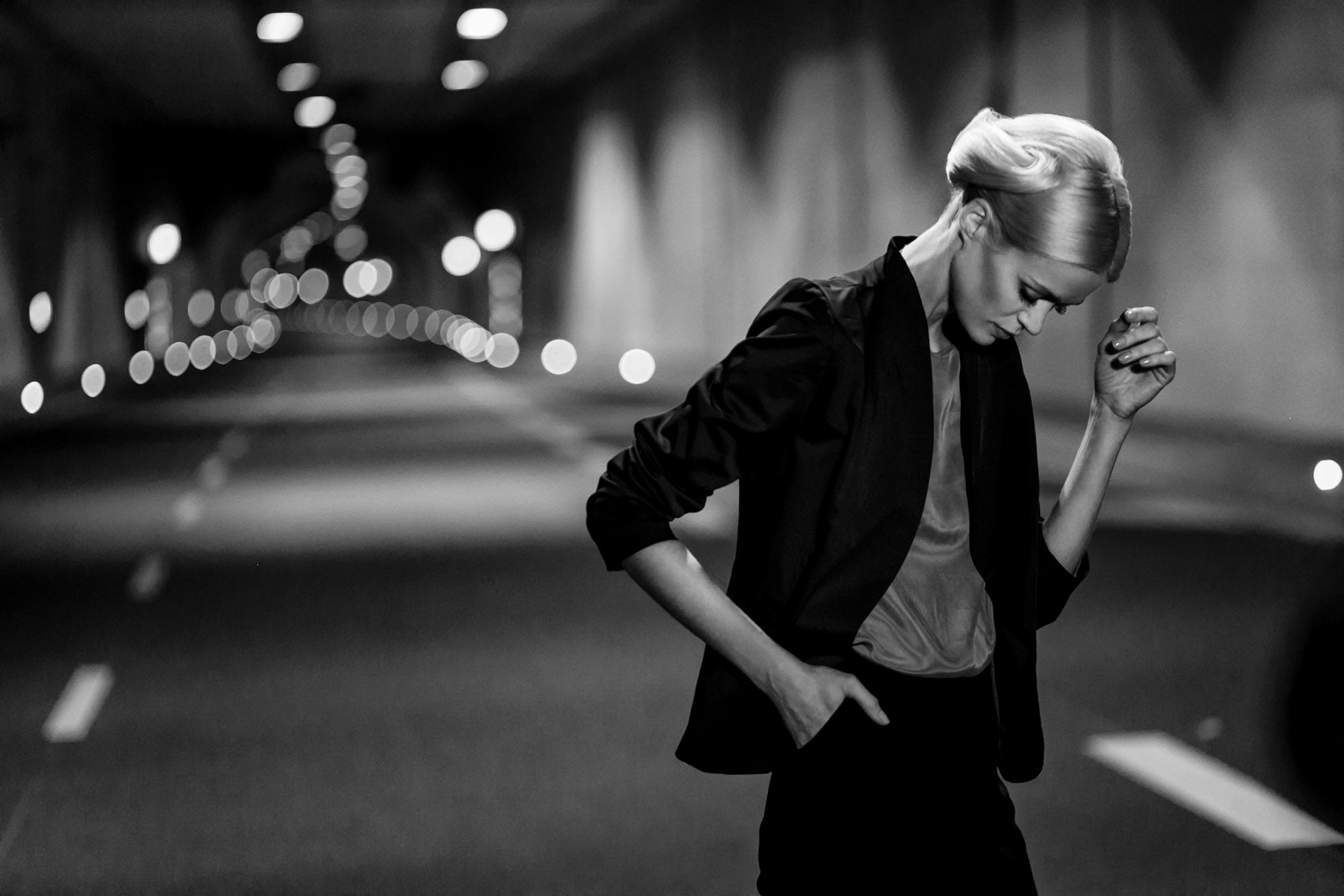 Czarno-białe zdjęcie uchwyciło stylową osobę w formalnym stroju, ucieleśniającą modową atmosferę edytorialną, gdy idzie samotnie w oświetlonym tunelu. Z elegancką fryzurą i jedną ręką w kieszeni, ich introspektywny wyraz uzupełnia rytmiczny wzór świateł za nimi.