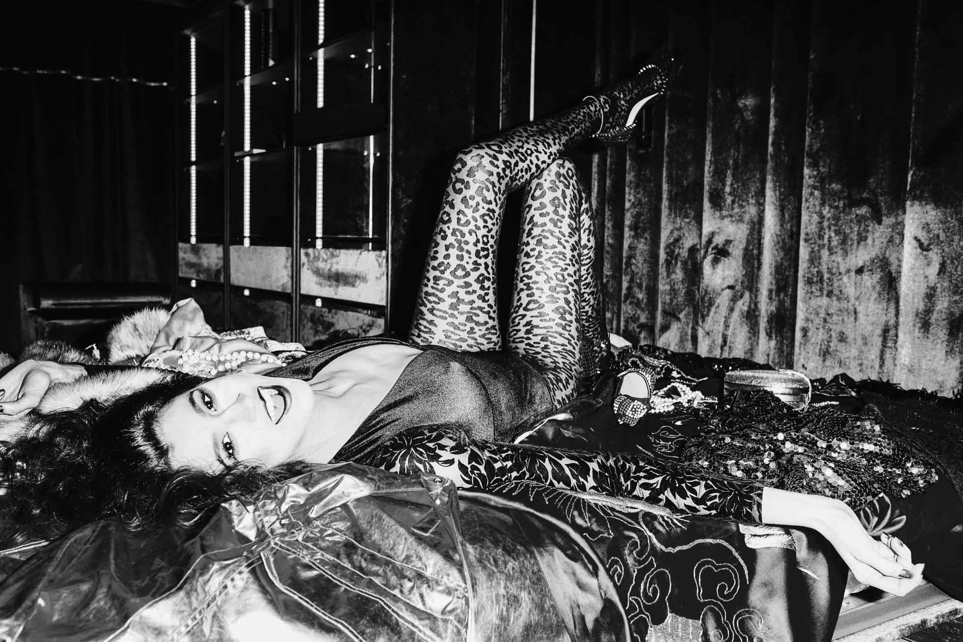 W słabo oświetlonym pomieszczeniu esencja edytorialu modowego ożywa, gdy osoba w odważnym makijażu i stroju w panterkę figlarnie wyleguje się na stosie ubrań. Jej nogi są uniesione w powietrzu, a żywa atmosfera została uchwycona w czarno-białej fotografii wizerunkowej.