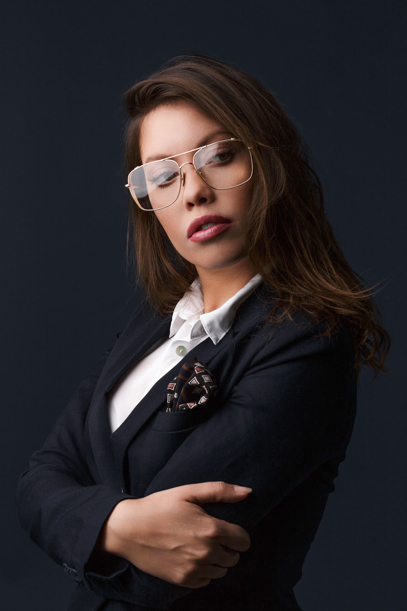 Osoba z długimi włosami i okularami, ubrana w białą koszulę i ciemny blezer, stoi ze skrzyżowanymi ramionami na ciemnym tle. Nosi ozdobną poszetkę w kieszeni blezera, co jest przykładem profesjonalnego wizerunku często uchwyconego w fotografia biznesowa i korporacyjna Trójmiasto.