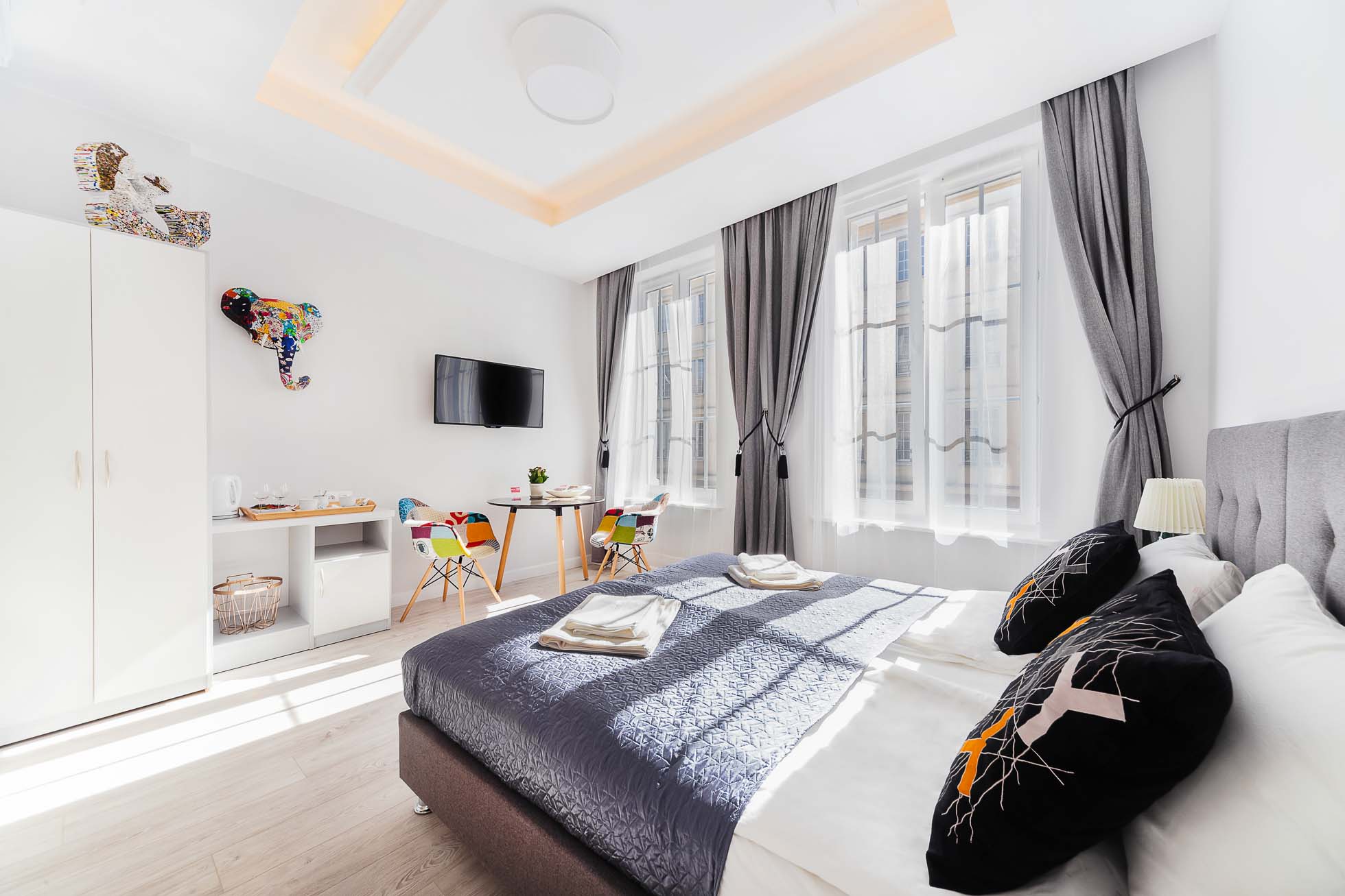 Jasny, nowoczesny pokój hotelowy wyposażony jest w starannie pościelone łóżko podwójne z ozdobnymi poduszkami, dwa duże okna z szarymi zasłonami, telewizor zamontowany na ścianie, białą szafę, mały stolik z kolorowymi krzesłami i fantazyjną dekorację ścienną przedstawiającą głowę słonia — idealną dla miłośników fotografii wnętrz trójmiasto.