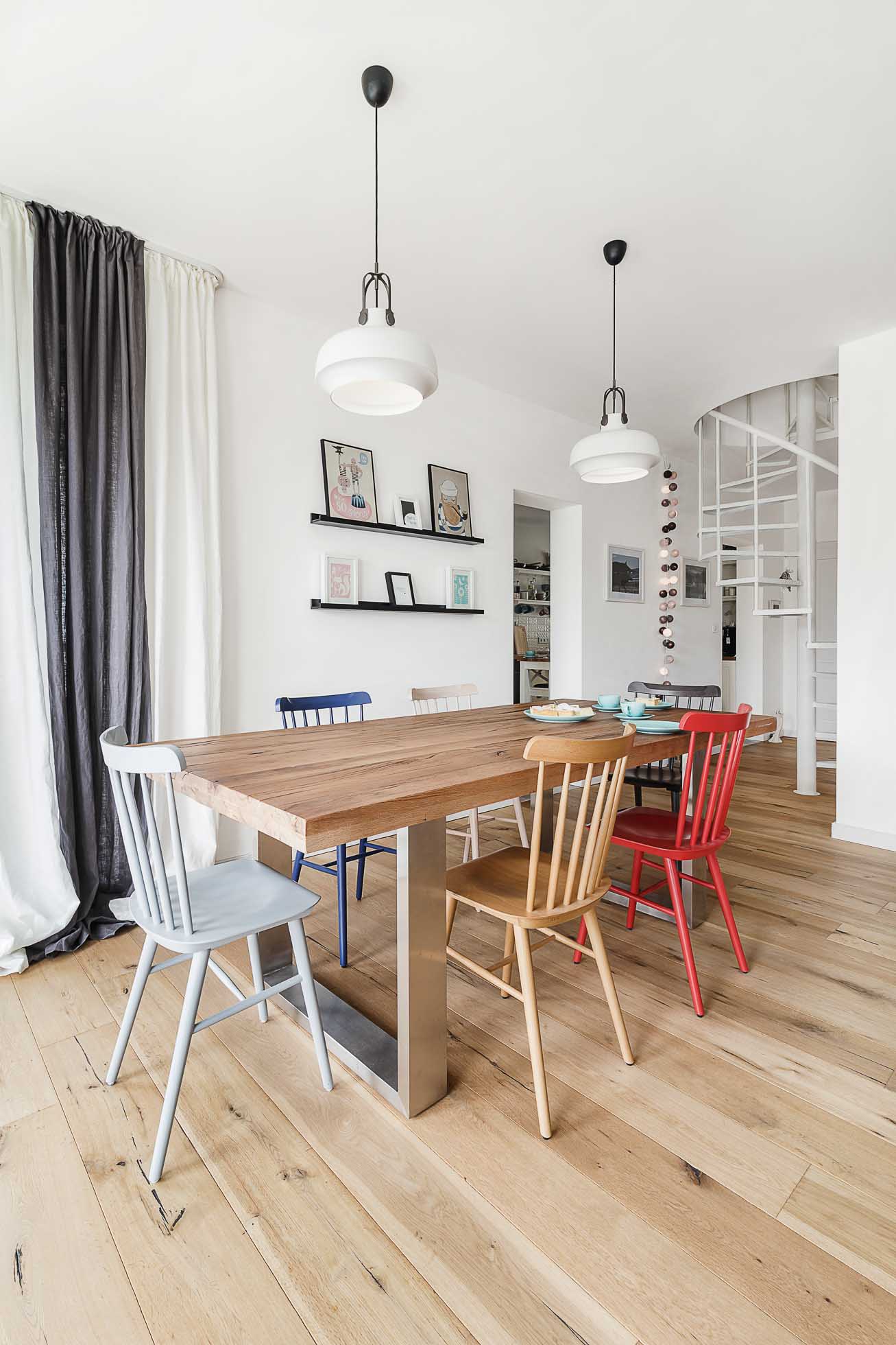 Nowoczesna jadalnia z drewnianym stołem i sześcioma krzesłami w różnych kolorach, uchwycona przez profesjonalnego fotografa mieszkań i wnetrz. Dwie wiszące lampy oświetlają stół. Czarno-biała zasłona częściowo zasłania okno po lewej stronie. Białe ściany eksponują dzieła sztuki, a w tle widać spiralne schody.