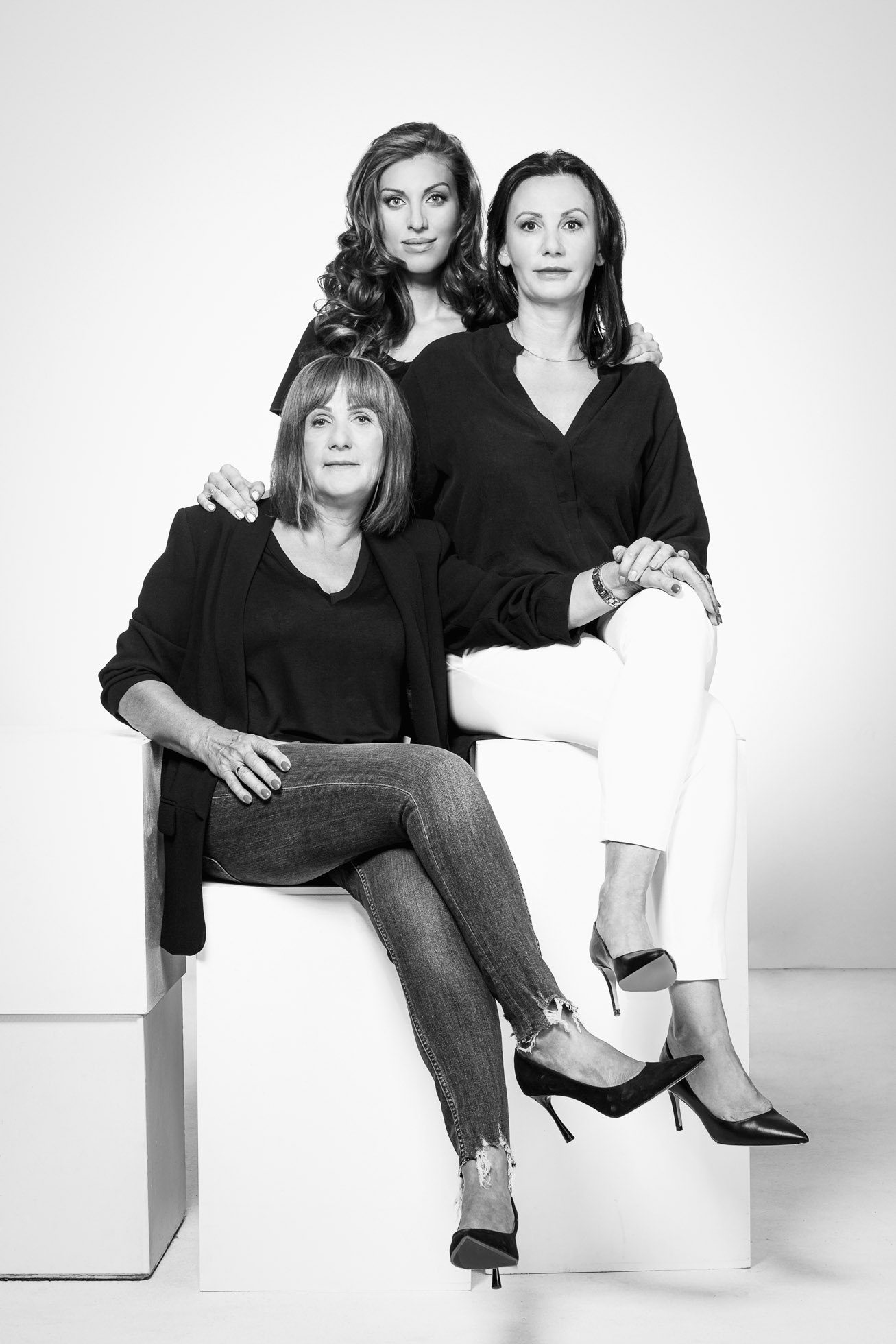 Czarno-biały portret trzech kobiet. Jedna z nich siedzi na niskim pudle, ubrana w marynarkę i dżinsy. Inna kobieta siedzi wyżej na białym sześcianie, ubrana w bluzkę i białe spodnie. Trzecia kobieta stoi za nimi, opierając ręce na ich ramionach, ubrana w bluzkę i spodnie. Profesjonalne sesje wizerunkowe Gdańsk doskonale oddają istotę.