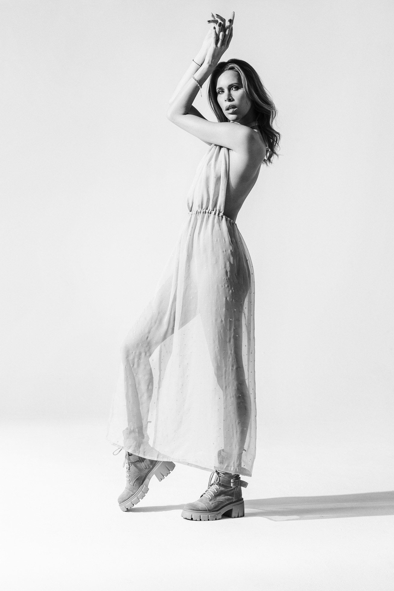Na zachwycającej czarno-białej fotografii wizerunkowej osoba pozuje elegancko w przezroczystej, zwiewnej sukience z dekoltem halter, w połączeniu z masywnymi butami. Zdjęcie podkreśla wdzięczną pozę i uderzający kontrast między delikatną tkaniną a solidnym obuwiem.
