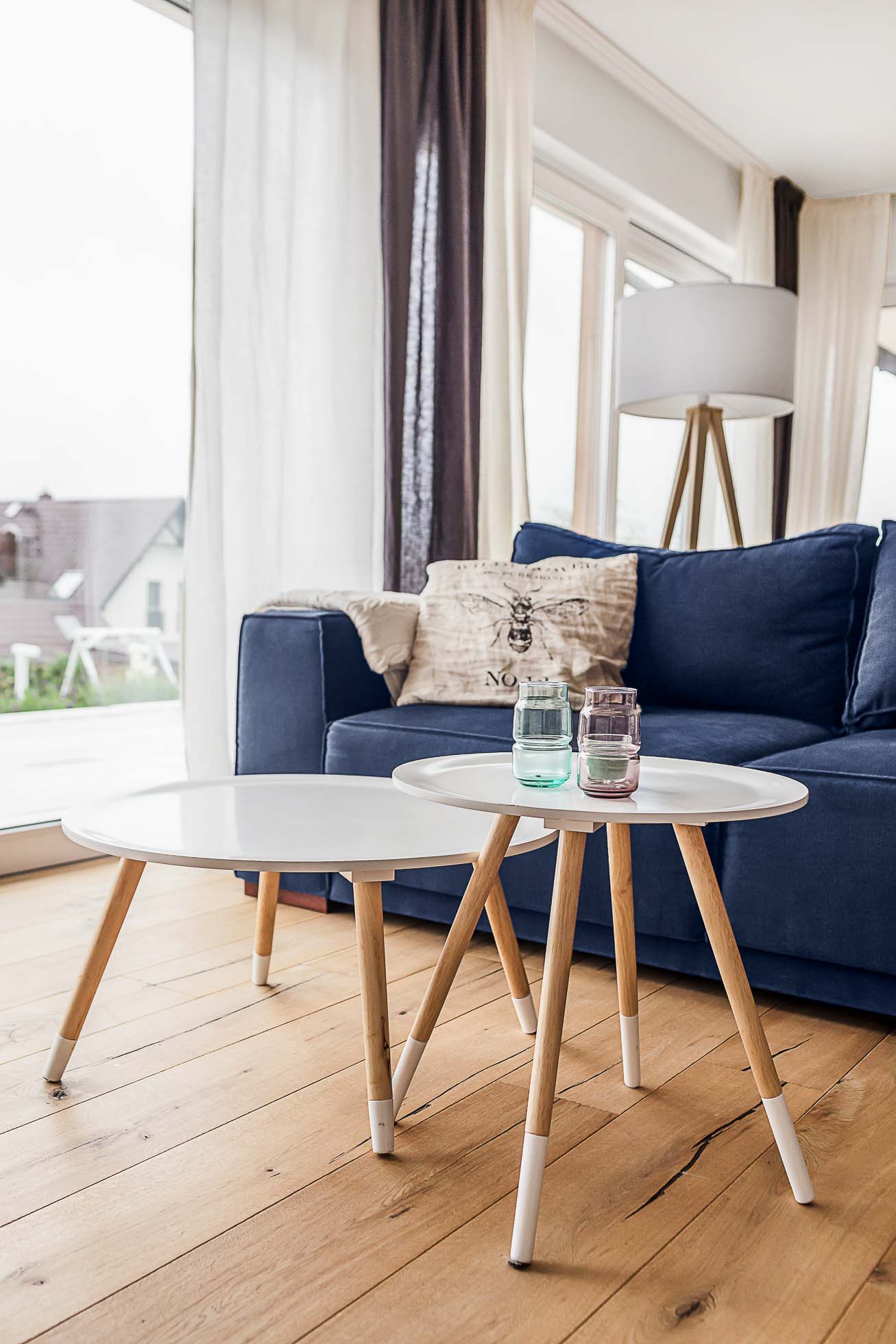 Nowoczesny salon z ciemnoniebieską sofą z białą poduszką, obok dwóch białych okrągłych stolików kawowych z drewnianymi nogami. Na stolikach znajdują się dwie małe świece w szklanych uchwytach. Pokój ma duże okna z białymi i szarymi zasłonami oraz drewnianą podłogę, idealną dla każdego Fotograf wnętrz premium w Gdańsku Gdyni i Sopocie.