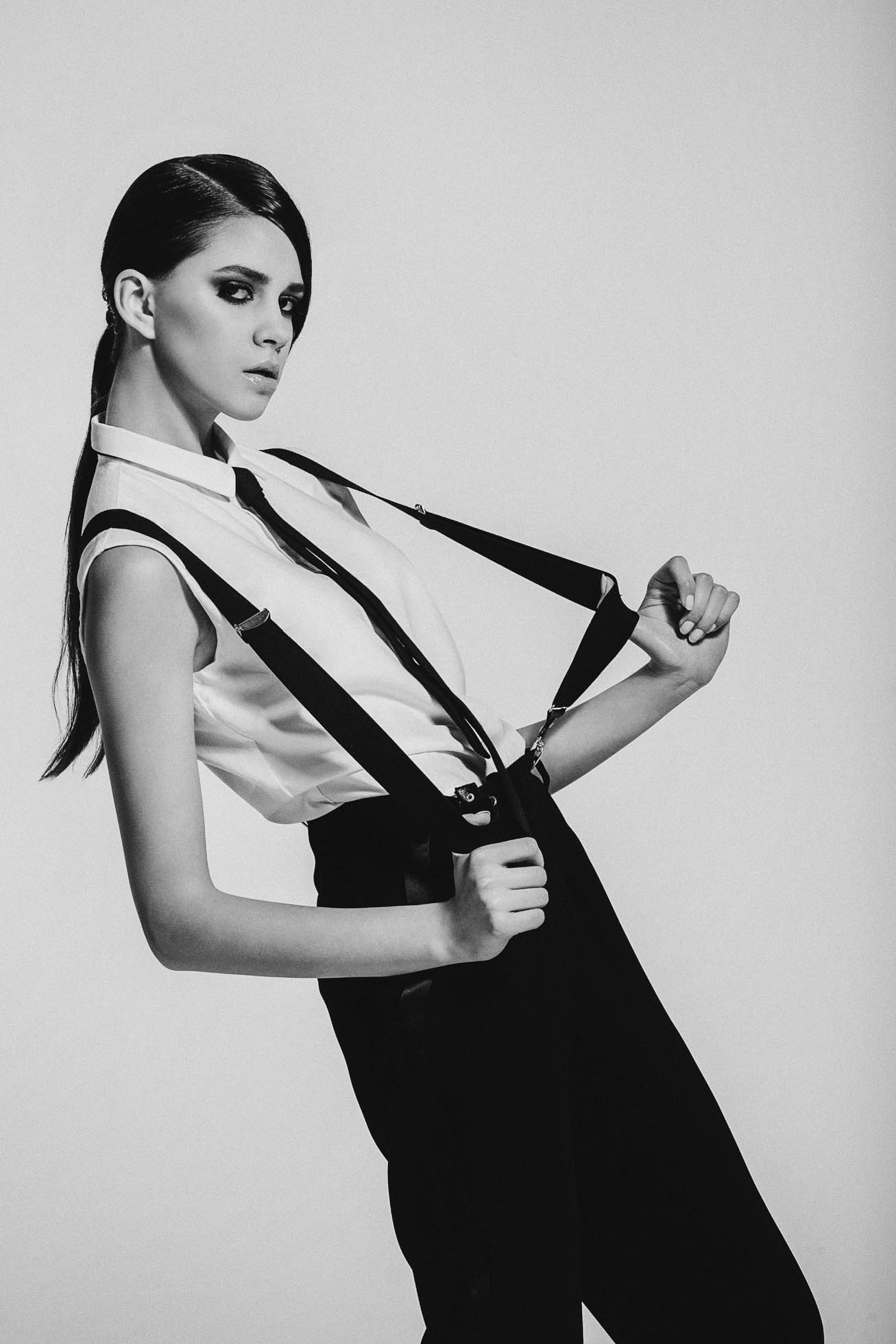 Kobieta emanuje pewnością siebie w monochromatycznej modzie, zakładając białą koszulkę bez rękawów i czarne szelki na czarne spodnie. Jej długie, ciemne włosy są związane, gdy wpatruje się uważnie w kamerę, uosabiając klasyczny styl edytorial na jednolitym tle.