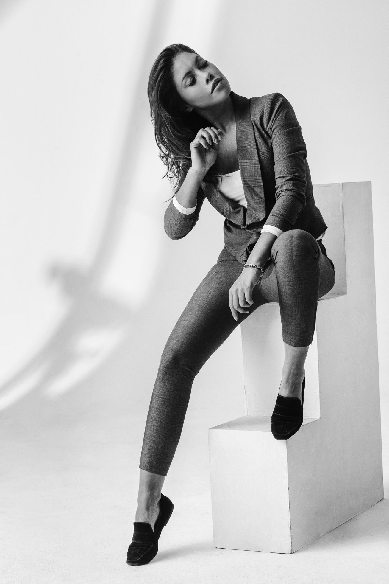 Kobieta siedzi na minimalistycznym, białym bloku, prezentując stylowy garnitur i mokasyny. Spogląda zamyślona w bok, ucieleśniając istotę modowego edytorialu fotografia wizerunkowa. Czarno-biały obraz podkreśla jej nowoczesny, wyrafinowany wygląd, uchwytując ponadczasową elegancję klimatu Sopotu.