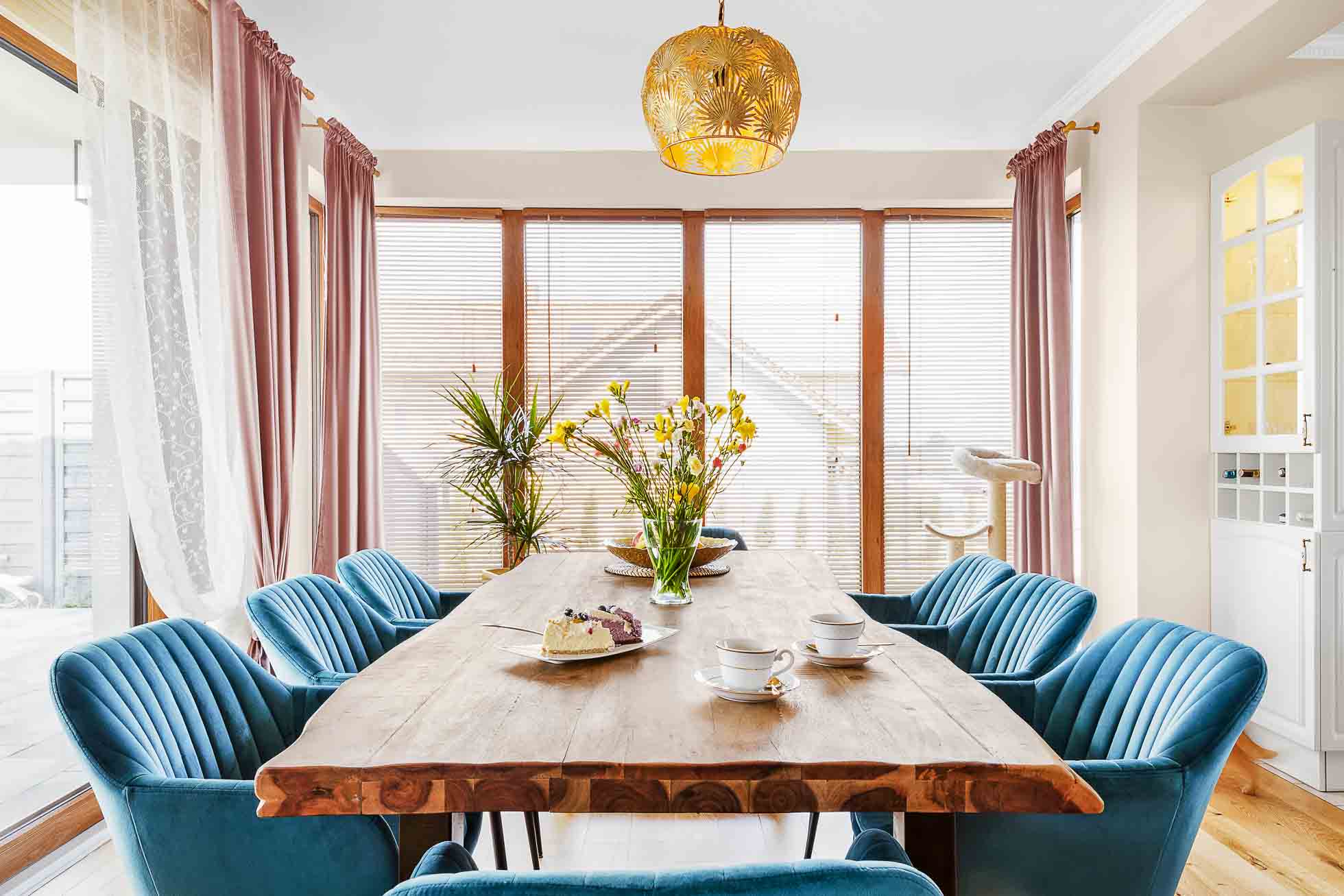 Jadalnia z drewnianym stołem otoczonym niebieskimi tapicerowanymi krzesłami. Na stole znajdują się dwie filiżanki do herbaty i talerze z wypiekami, a na środku stoi wazon z żółtymi kwiatami. Duże okna z żaluzjami i firankami w tle, pod złotą wiszącą lampą, idealne do fotografia wnętrz Trójmiasto – zdjęcia apartamentów i lokali komercyjnych.
