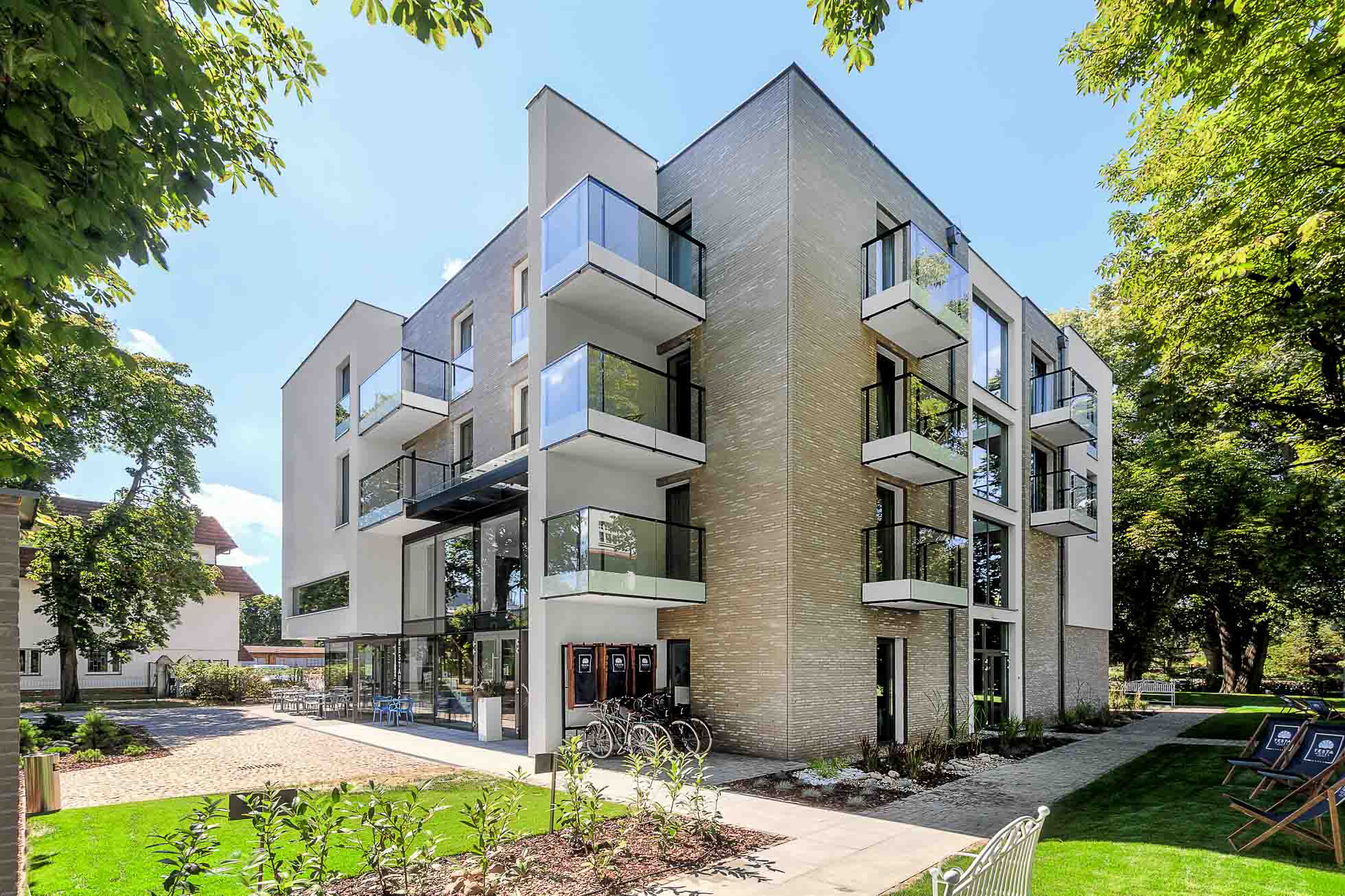 Nowoczesny czteropiętrowy budynek apartamentowy ze szklanymi balkonami i eleganckim, współczesnym designem. Budynek położony jest wśród zagospodarowanego ogrodu z drzewami i zadbanymi trawnikami. Są tam patio i miejsca do siedzenia, zapewniając mieszkańcom spokojną przestrzeń na zewnątrz. Idealny dla fotografia wnętrz Trójmiasto – zdjęcia apartamentów i lokali komercyjnych.