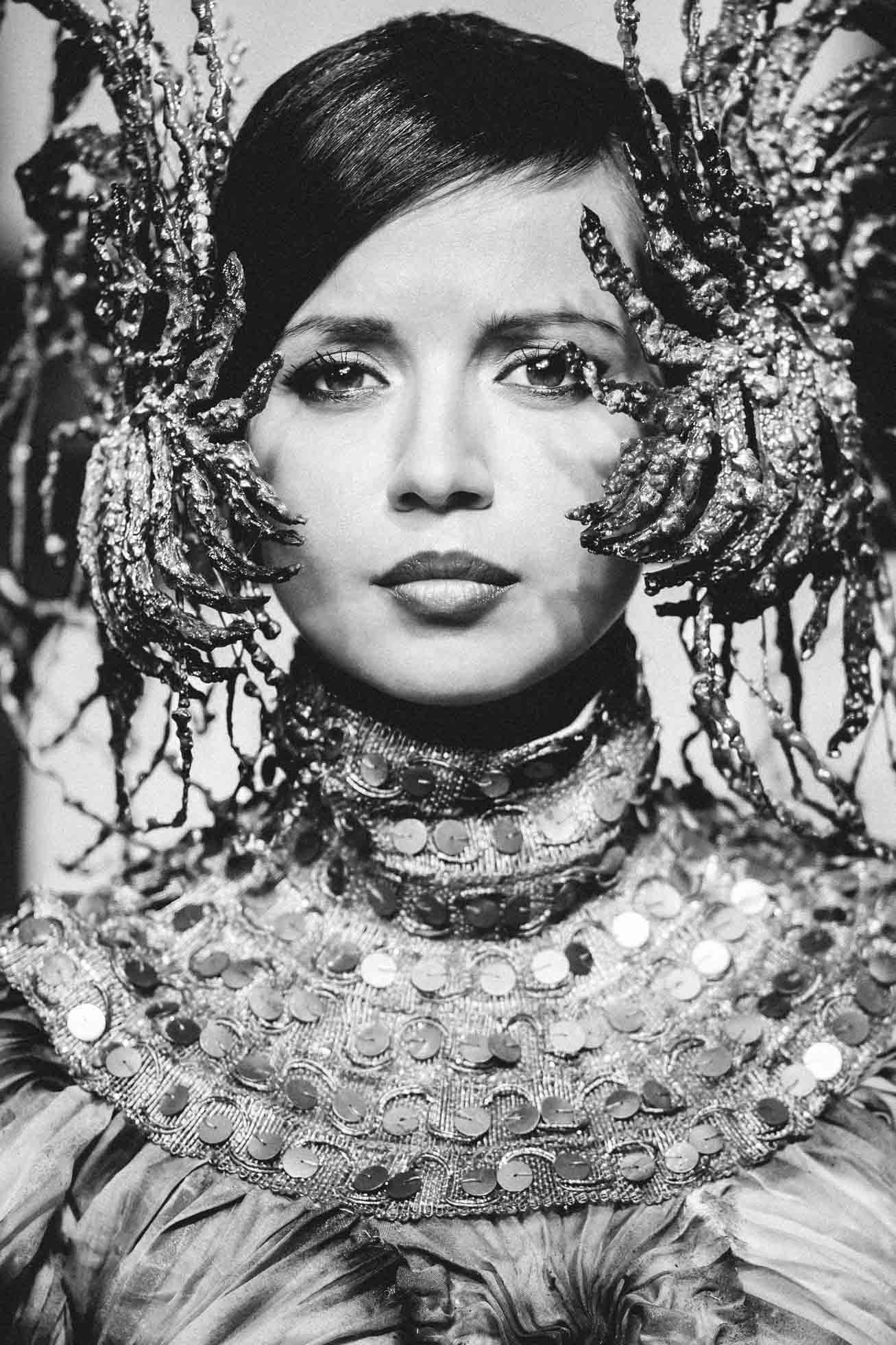 Kobieta patrzy przed siebie, mając na sobie misterne i ozdobne nakrycie głowy z metalowymi przedłużeniami. Jej strój z wysokim kołnierzem zdobią błyszczące okrągłe ozdoby, tworząc królewski i artystyczny wygląd. Ta oszałamiająca fotografia edytorialowa z Trójmiasta uchwyciła jej elegancję w czerni i bieli.