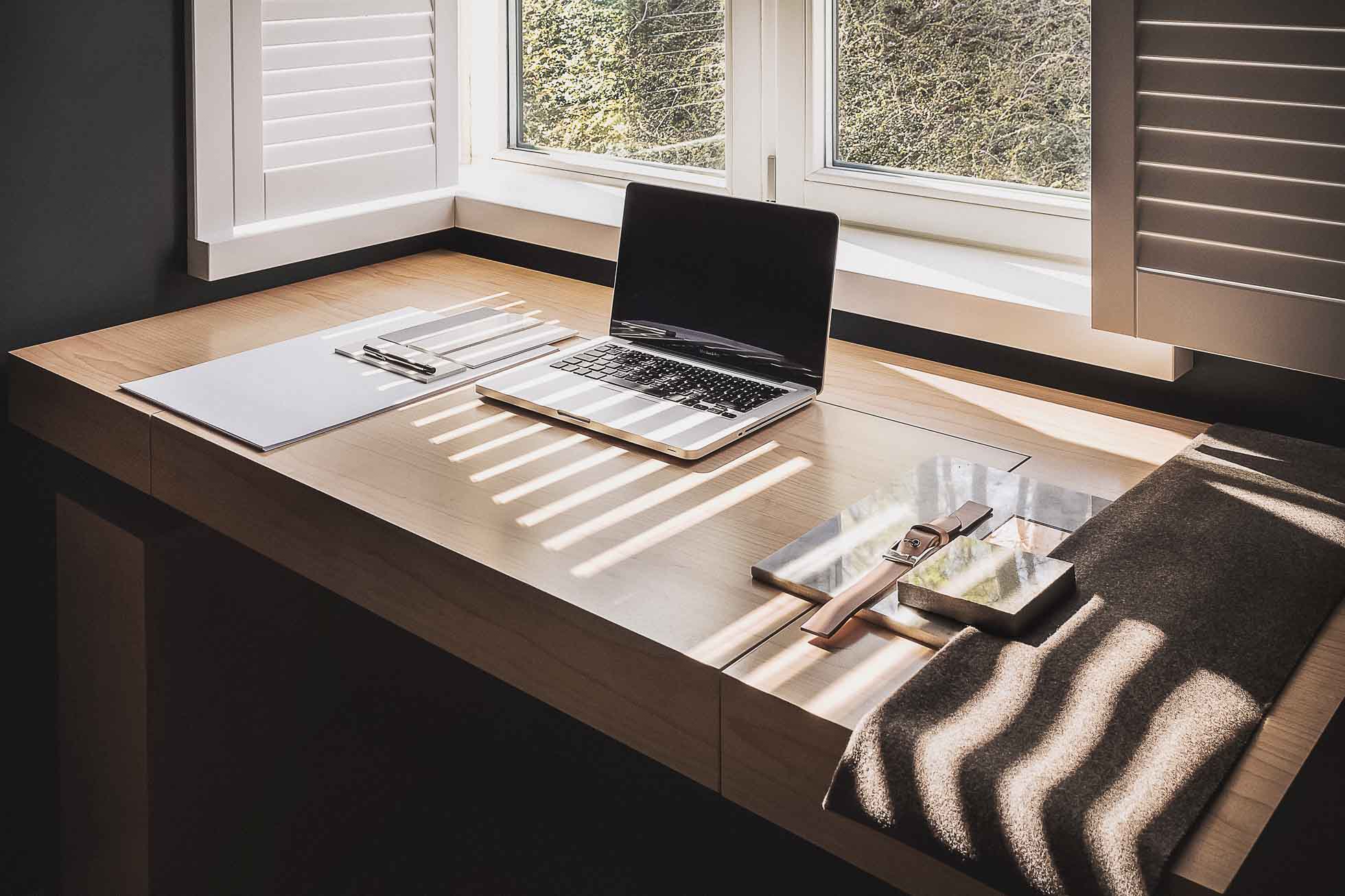 Minimalistyczne miejsce pracy z drewnianym biurkiem umieszczonym pod oknem z białymi okiennicami, częściowo otwartym, aby wpuścić światło słoneczne. Na biurku znajduje się otwarty laptop, notatnik z długopisem i książka w oprawie materiałowej. Idealne dla Fotografia architektury Trójmiasto – zdjęcia wnętrz i budynków, emanuje spokojem i produktywnością.