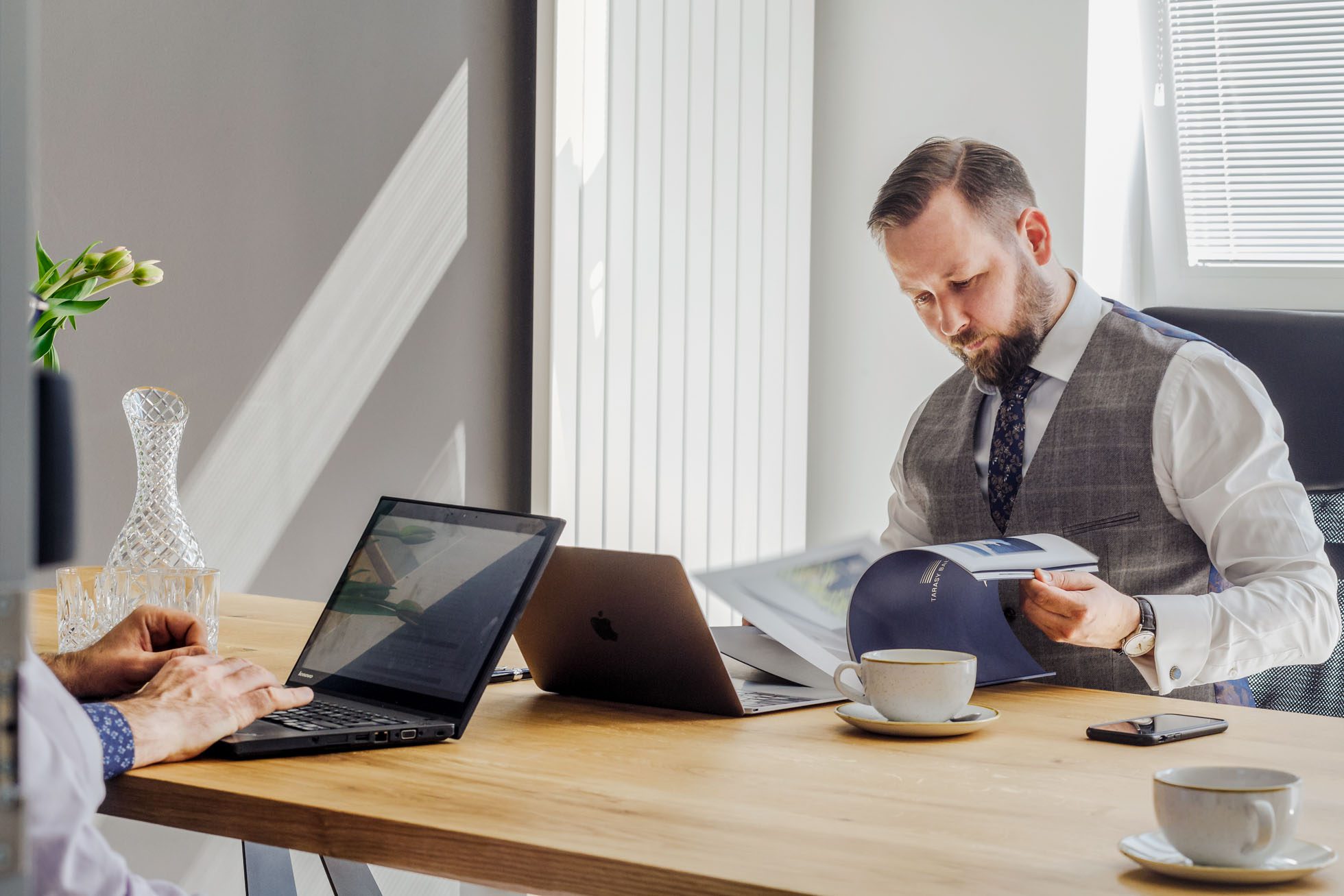 Dwóch mężczyzn pracuje przy drewnianym biurku w jasnym, słonecznym biurze. Jeden z nich skupia się na laptopie, podczas gdy drugi przegląda dokumenty. Dwa laptopy są otwarte na stole obok kubków do kawy i smartfona. Ustawienie sugeruje kreatywne środowisko typowe dla Fotografii biznesowej i korporacyjnej Trójmiasto.