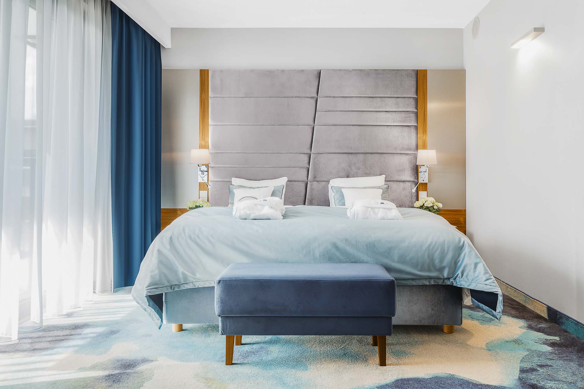 Nowoczesna sypialnia hotelowa z dużym łóżkiem z szarym tapicerowanym wezgłowiem i starannie ułożoną jasnoniebieską pościelą. Obok łóżka znajdują się lampy ścienne i małe stoliki nocne z białymi kwiatami. Niebieska ława znajduje się u stóp łóżka, a podłogę pokrywa wzorzysty dywan. Idealne dla Fotografia wnętrz Trójmiasto – zdjęcia apartamentów i lokali komercyjnych.