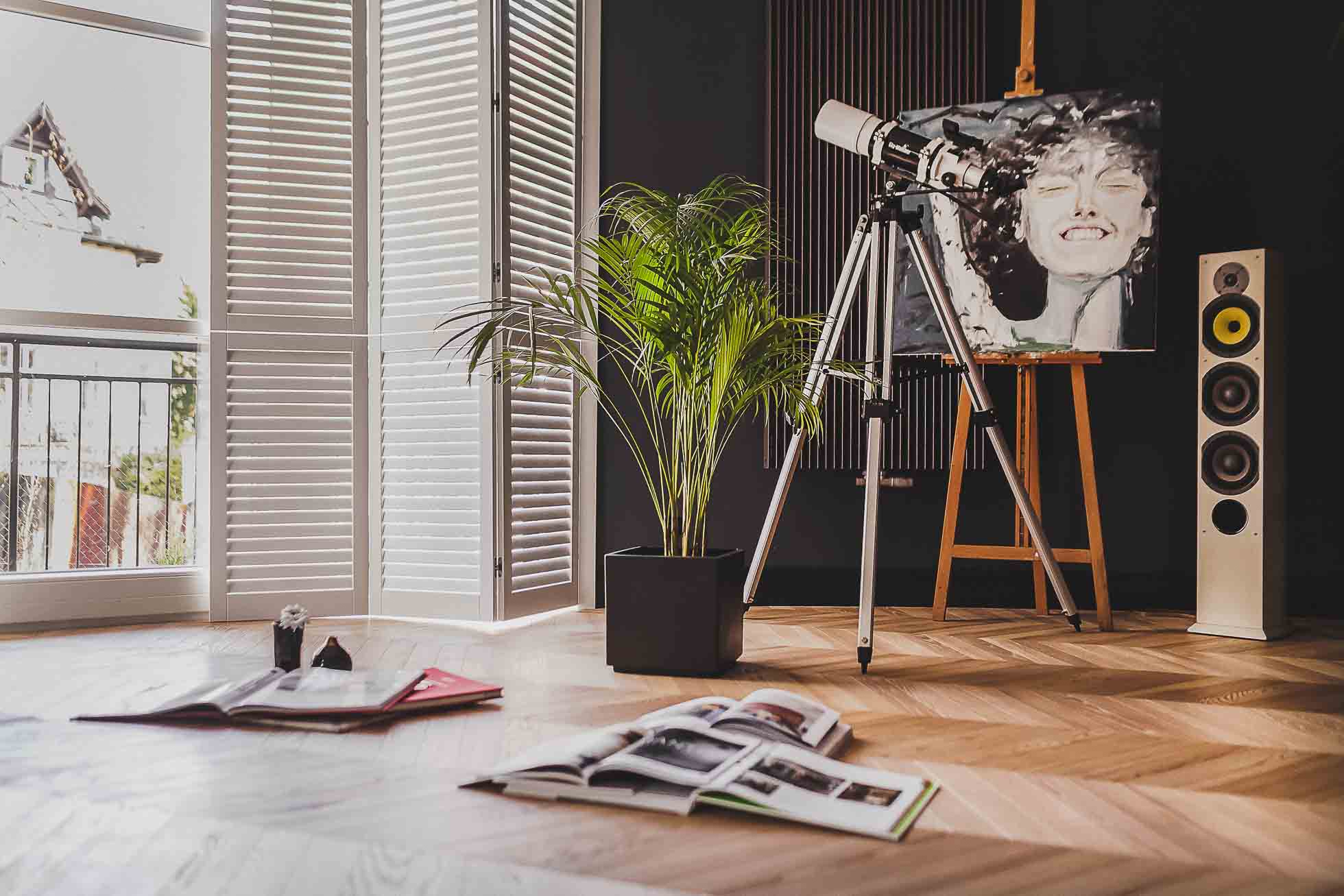 Dobrze oświetlony pokój z drewnianą podłogą zawiera sztalugę z czarno-białym portretem, dużą roślinę doniczkową, teleskop i głośnik stojący na podłodze. Otwarte książki i mała roślina leżą na podłodze. Duże okna z białymi okiennicami wypełniają tło, idealne do projektów *Fotografia wnętrz Trójmiasto*.