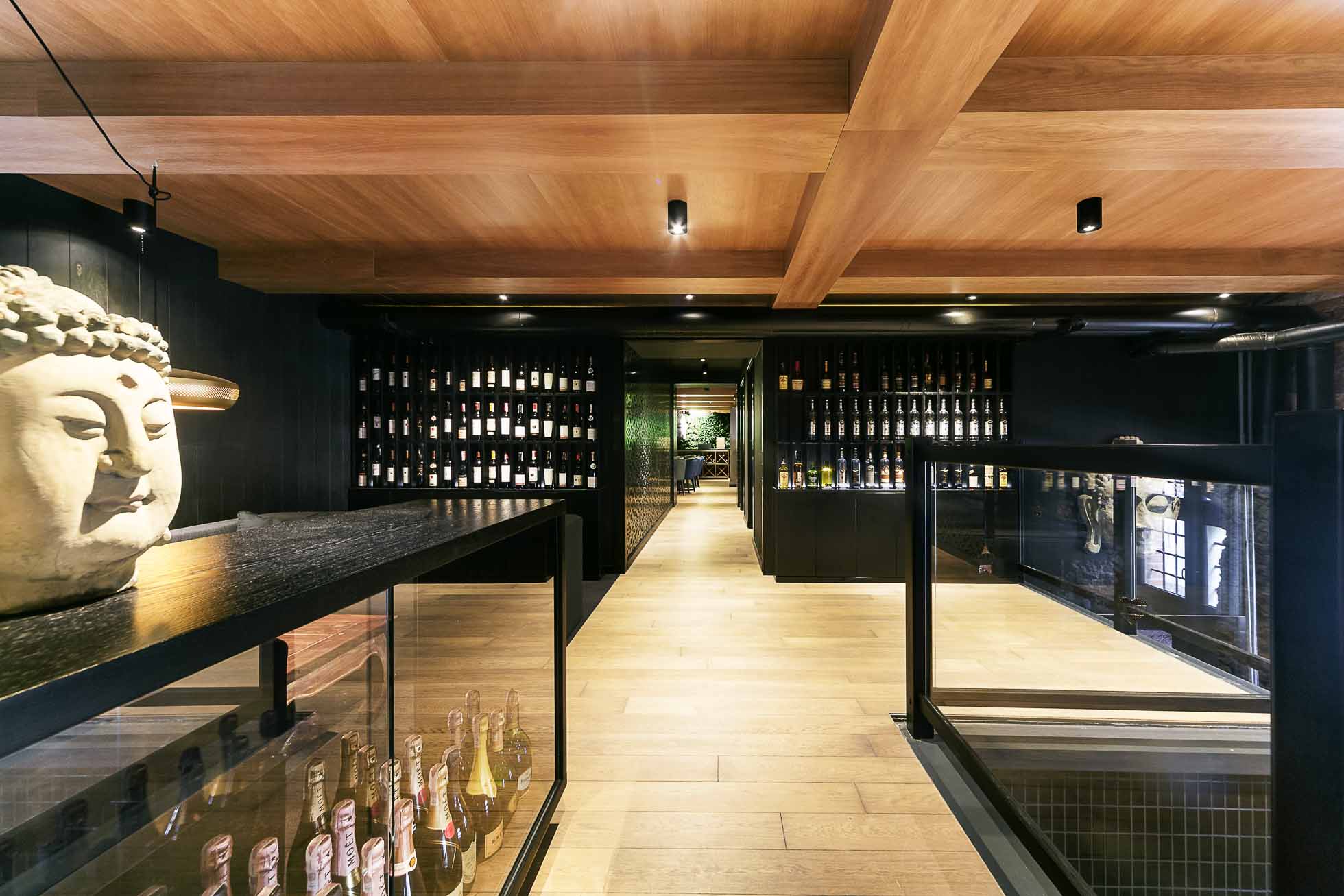 Nowoczesne wnętrze tajskiej restauracji z drewnianym sufitem i podłogą, z półkami wypełnionymi butelkami wina. Duża statua głowy Buddy umieszczona na ladzie na pierwszym planie. Oświetlenie jest ciepłe, tworząc przytulną atmosferę. Sesje fotograficzne wnętrz Gdańsk Gdynia Sopot – usługi fotograficzne doskonale podkreślają nastrój.