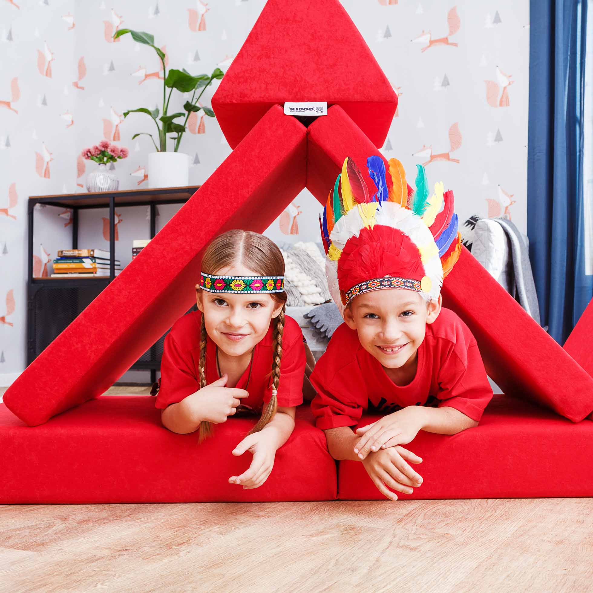 Dwoje dzieci bawi się na czerwonym forcie z poduszek, oboje z długimi warkoczami i w czerwonych koszulkach. Dziecko po prawej ma na głowie kolorowy pióropusz, a to po lewej ma opaskę z koralików. W tle mały stolik z roślinami dodaje uroku. Idealne do fotografia komercyjna Trójmiasto.
