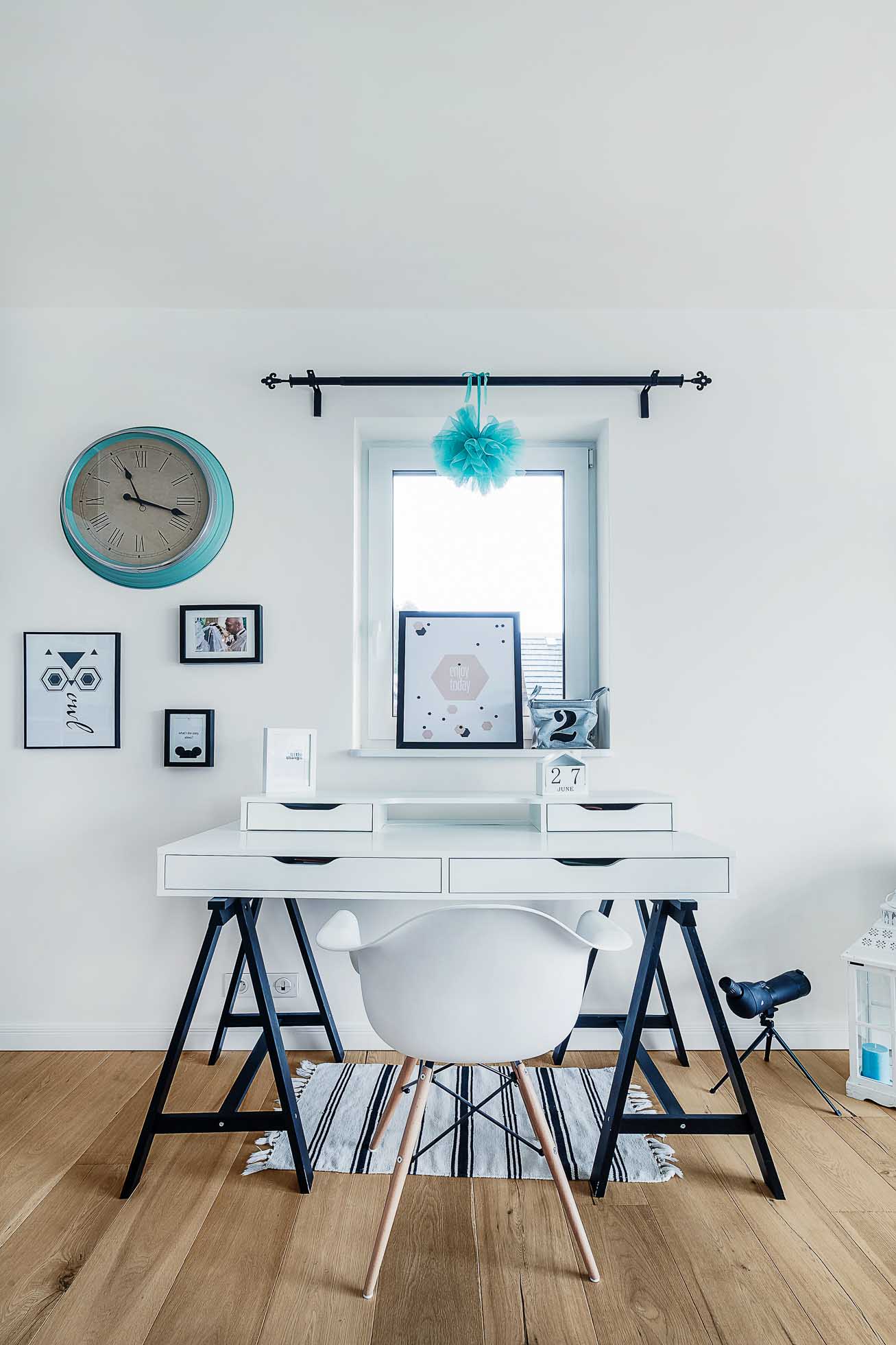 Nowoczesne, minimalistyczne miejsce pracy z białym biurkiem z czarnymi nogami, białym krzesłem z drewnianymi nogami, małym zestawem szuflad, oprawionymi dziełami sztuki i zegarem na ścianie. Okno z niebieskim pomponem znajduje się centralnie nad biurkiem, co pozwala na wpadanie naturalnego światła — idealne dla fotografia wnętrz Trójmiasto.