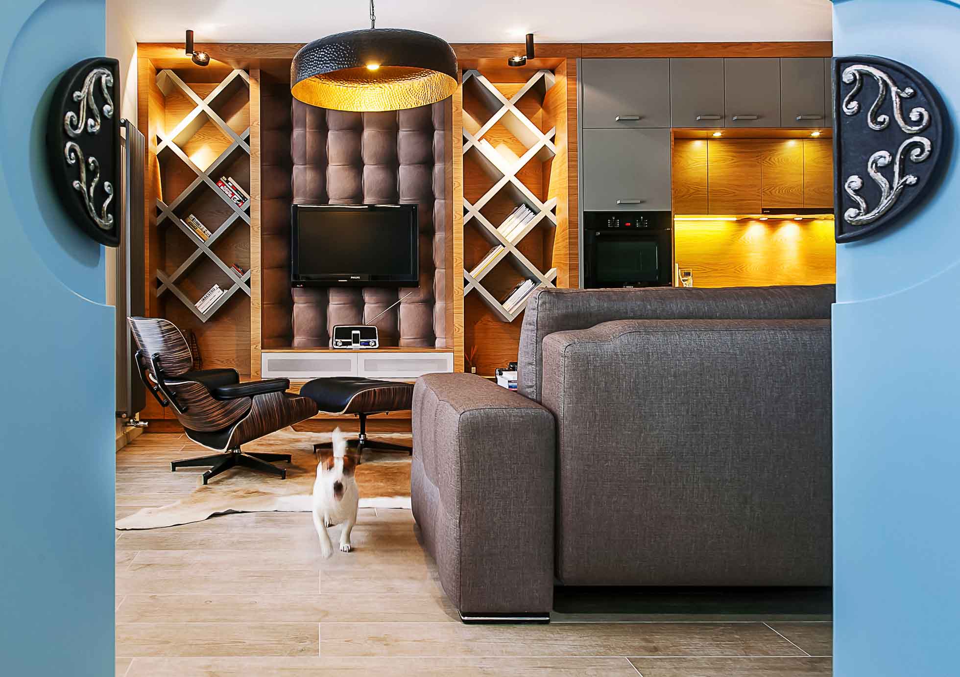 Nowoczesny salon z półkami w kształcie litery X wypełnionymi książkami, pikowaną ścianą z zamontowanym telewizorem, sofą, fotelem i lampą podłogową. Pies idzie w stronę aparatu po drewnianej podłodze. Przestrzeń jest ciepło oświetlona oświetleniem wnękowym — idealnym do fotografia wnętrz Trójmiasto.