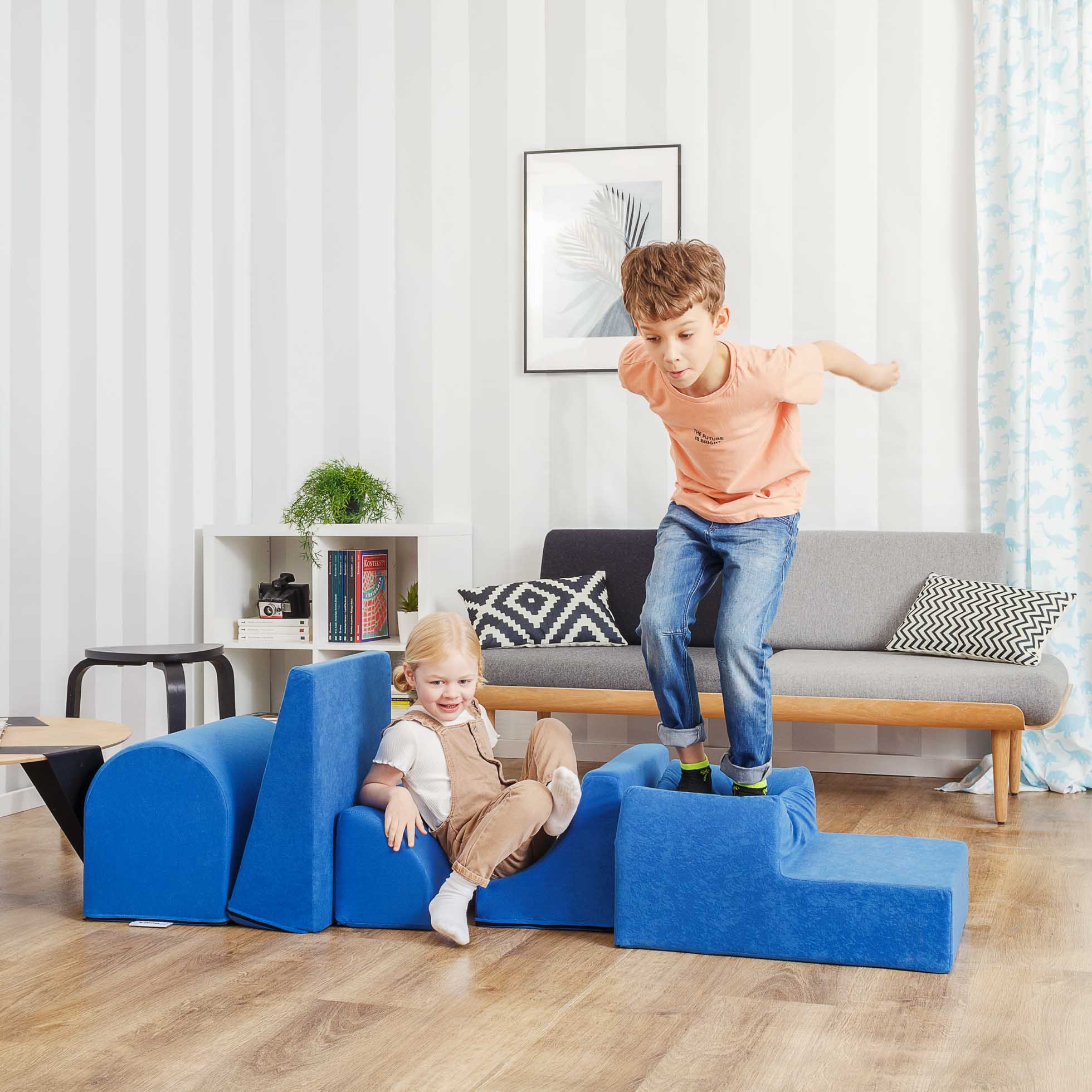 Dwoje dzieci bawiących się w jasnym, nowoczesnym salonie. Jedno dziecko, ubrane w pomarańczową koszulę i dżinsy, zeskakuje z modułowej niebieskiej konstrukcji z pianki. Drugie, siedzące na konstrukcji, ubrane w białą koszulę i brązowe spodnie, uśmiecha się. W tle sofa i regał na książki — idealne dla Fotografia reklamowa Trójmiasto.