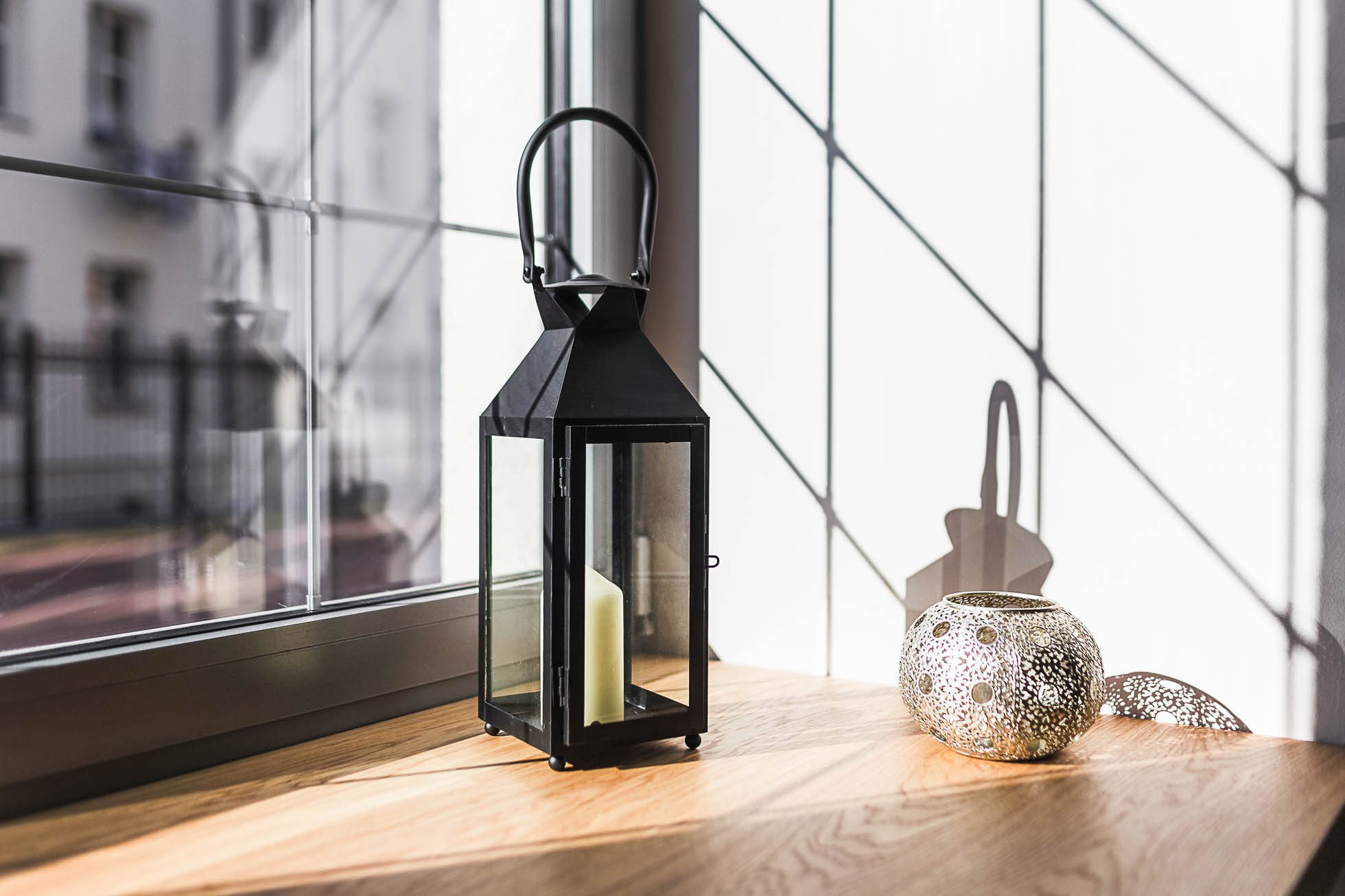 Czarna latarnia z białą świecą w środku stoi na drewnianym parapecie, obok ozdobnego srebrnego świecznika. Światło słoneczne wpada przez okno, rzucając cienie na drewnianą powierzchnię i ścianę. W pomieszczeniu panuje jasna i przestronna atmosfera — idealna do fotografia architektury Trójmiasto – zdjęcia wnętrz i budynków.