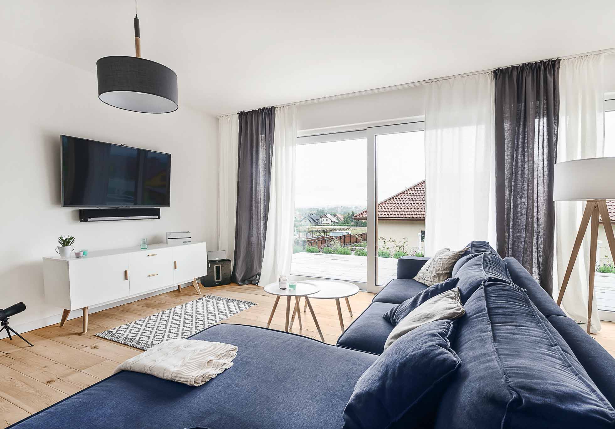 Nowoczesny salon z dużą niebieską sofą modułową naprzeciwko zamontowanego na ścianie telewizora z płaskim ekranem. W pokoju znajduje się biała konsola multimedialna, okrągły stolik kawowy, wysoka lampa podłogowa i duże okna z cienkimi firankami wpuszczającymi naturalne światło. Jasne drewniane podłogi dopełniają przestrzeń. Sesje fotograficzne wnętrz Gdańsk Gdynia Sopot – usługi fotograficzne.